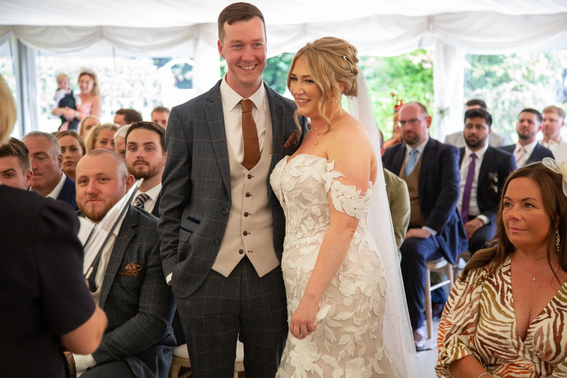 A smiling bride and groom stand together during their wedding ceremony inside a bright white marquee, surrounded by seated guests.