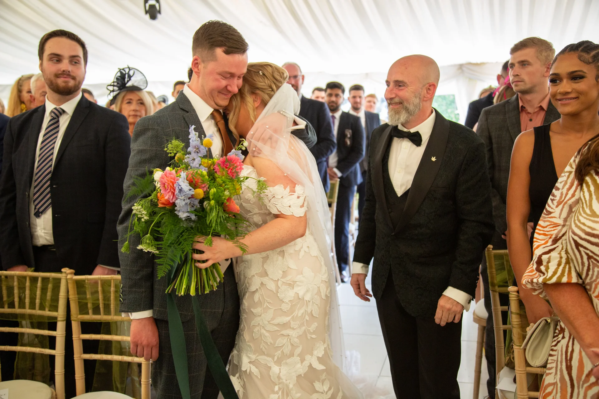 A groom gently kisses the bride's forehead as they embrace during their marquee wedding ceremony, surrounded by guests.