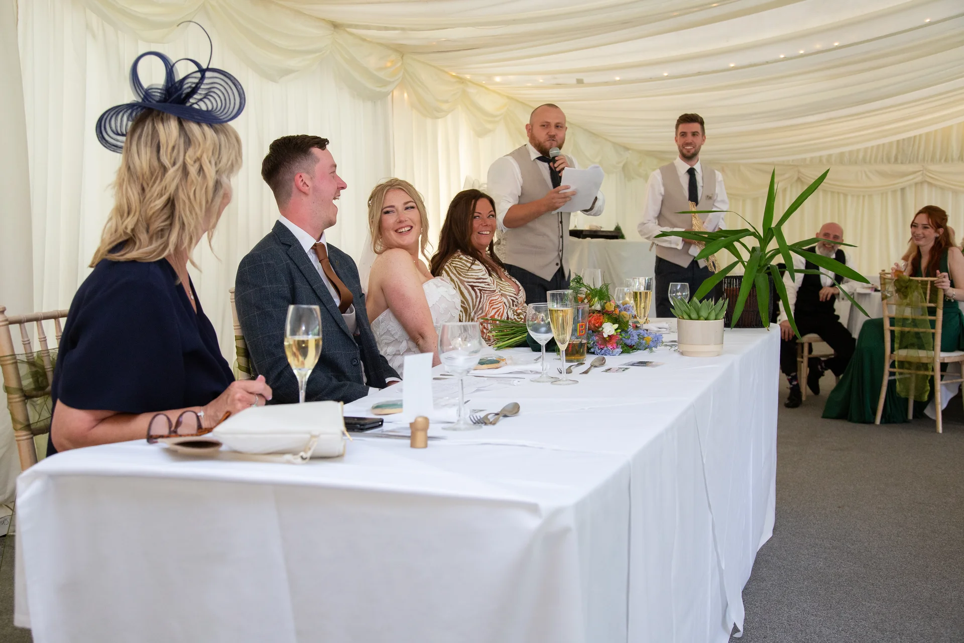 The newlywed couple and wedding guests seated at the top table in a draped marquee laughing during the wedding speeches.