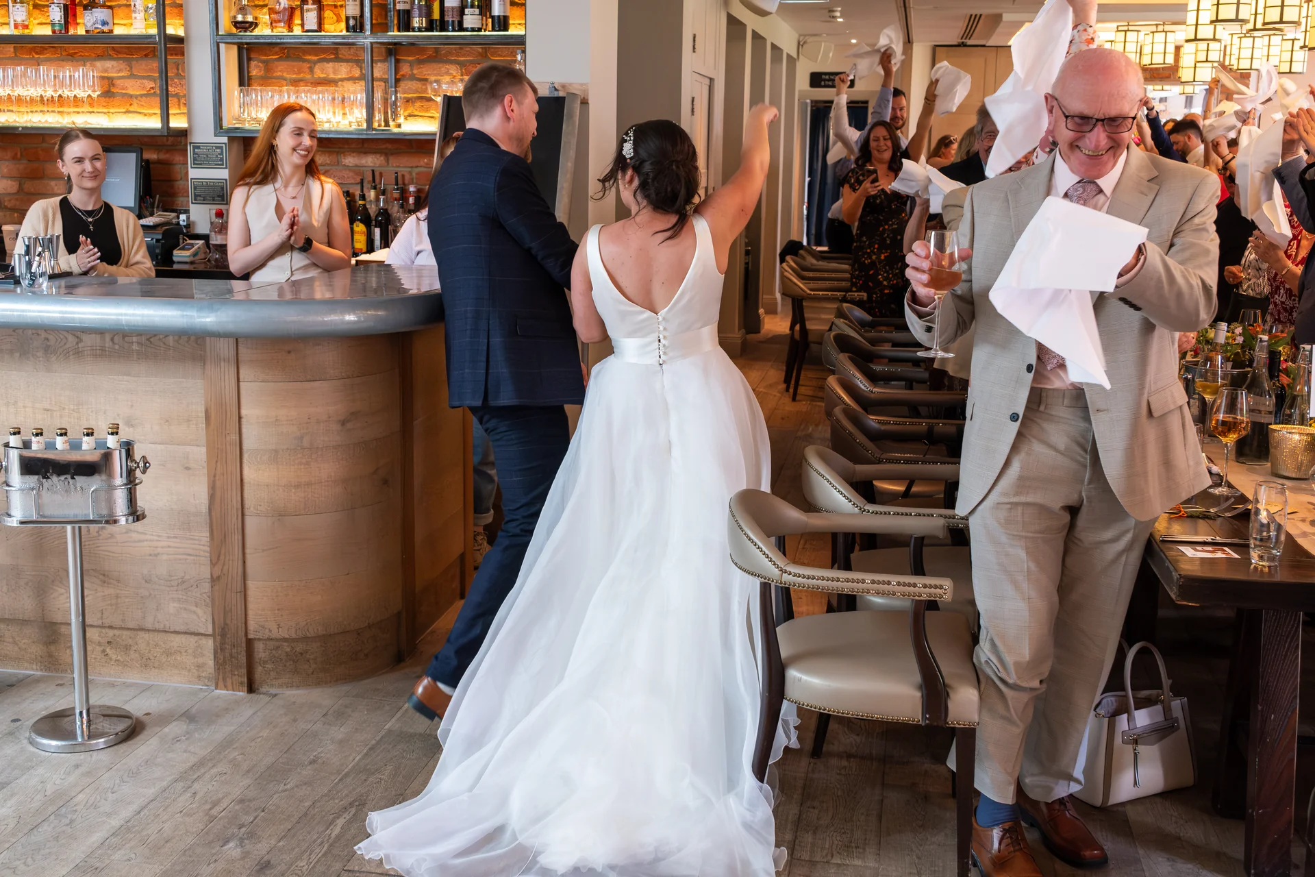 A joyful wedding reception in a stylish pub where guests wave their napkins in celebration as the bride in a classic white gown and the groom walk past tables.
