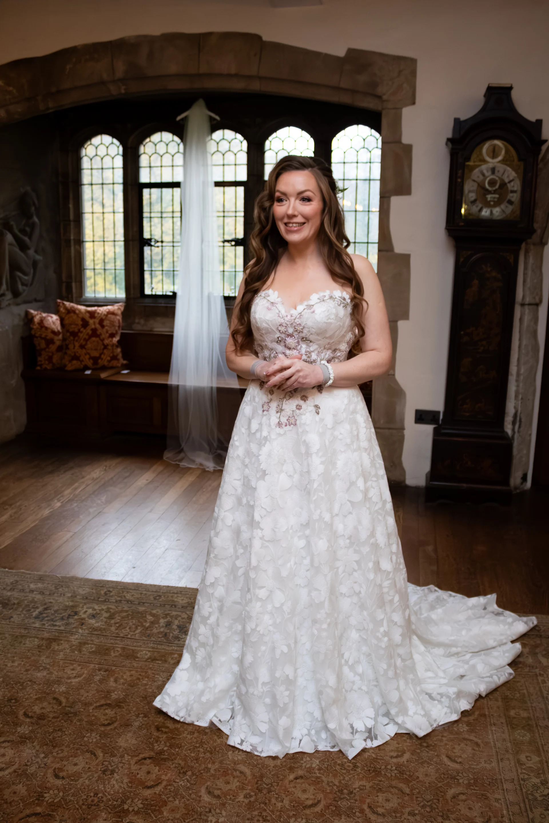 A smiling bride in an A-line gown with floral lace applique stands inside a historic manor house with stone archways and lead glass windows.