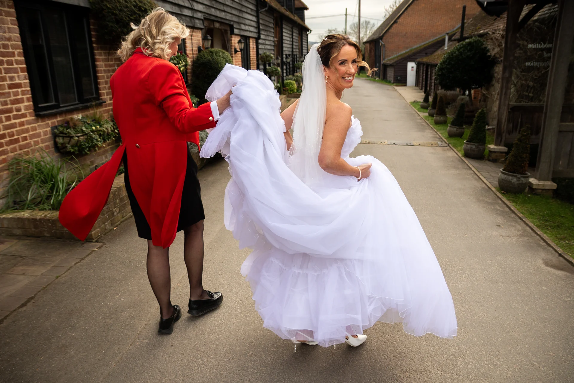 A smiling bride wearing a white a-line wedding dress and veil looks over her shoulder while walking down a pathway as someone assists with her dress.