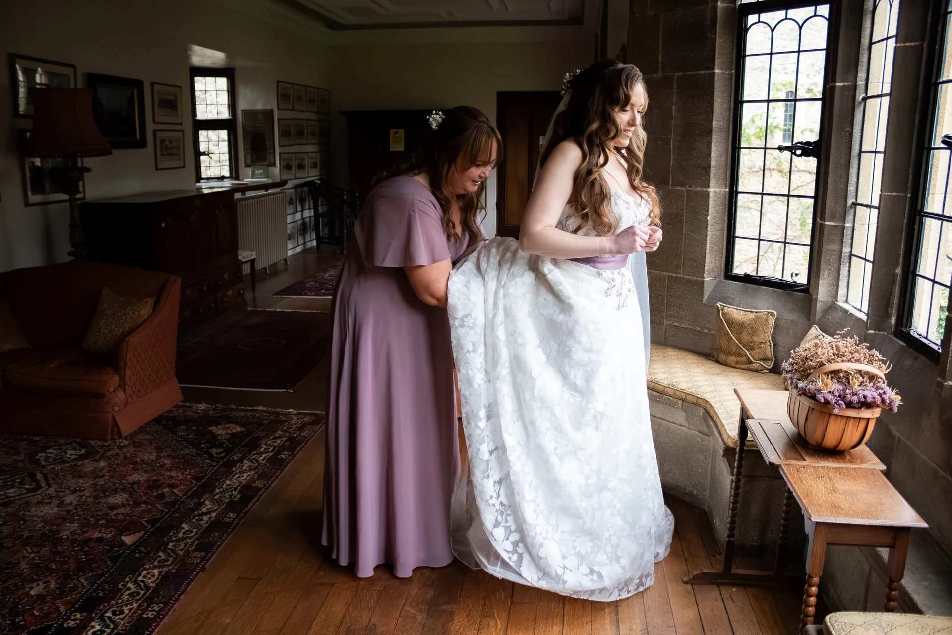 A bridesmaid in a purple dress assists the bride with her floral-patterned lace gown near a stone-mullioned window in a historic manor house.