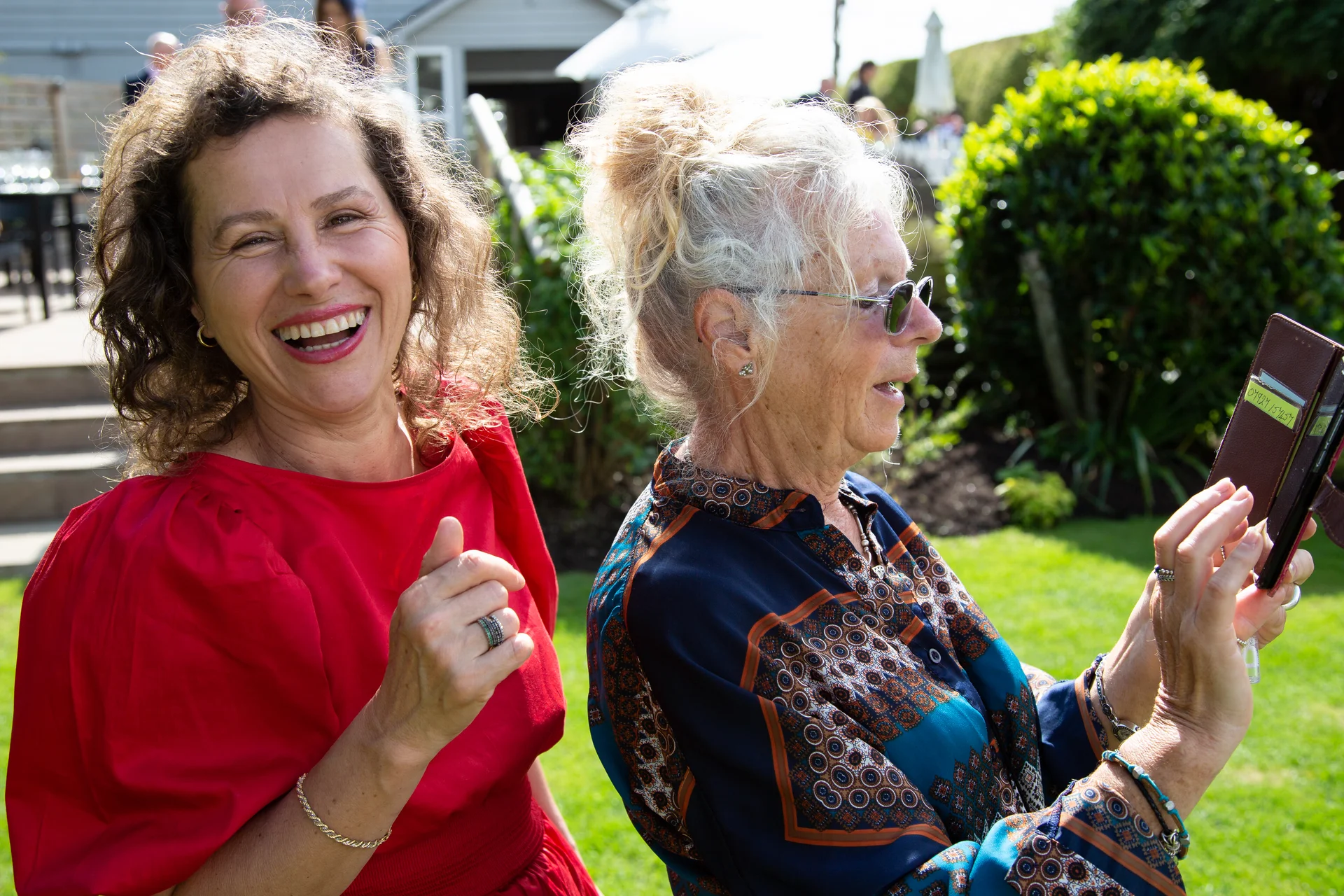 Two smiling women in bright outfits enjoying an outdoor wedding celebration while one holds up a mobile phone to take a picture.