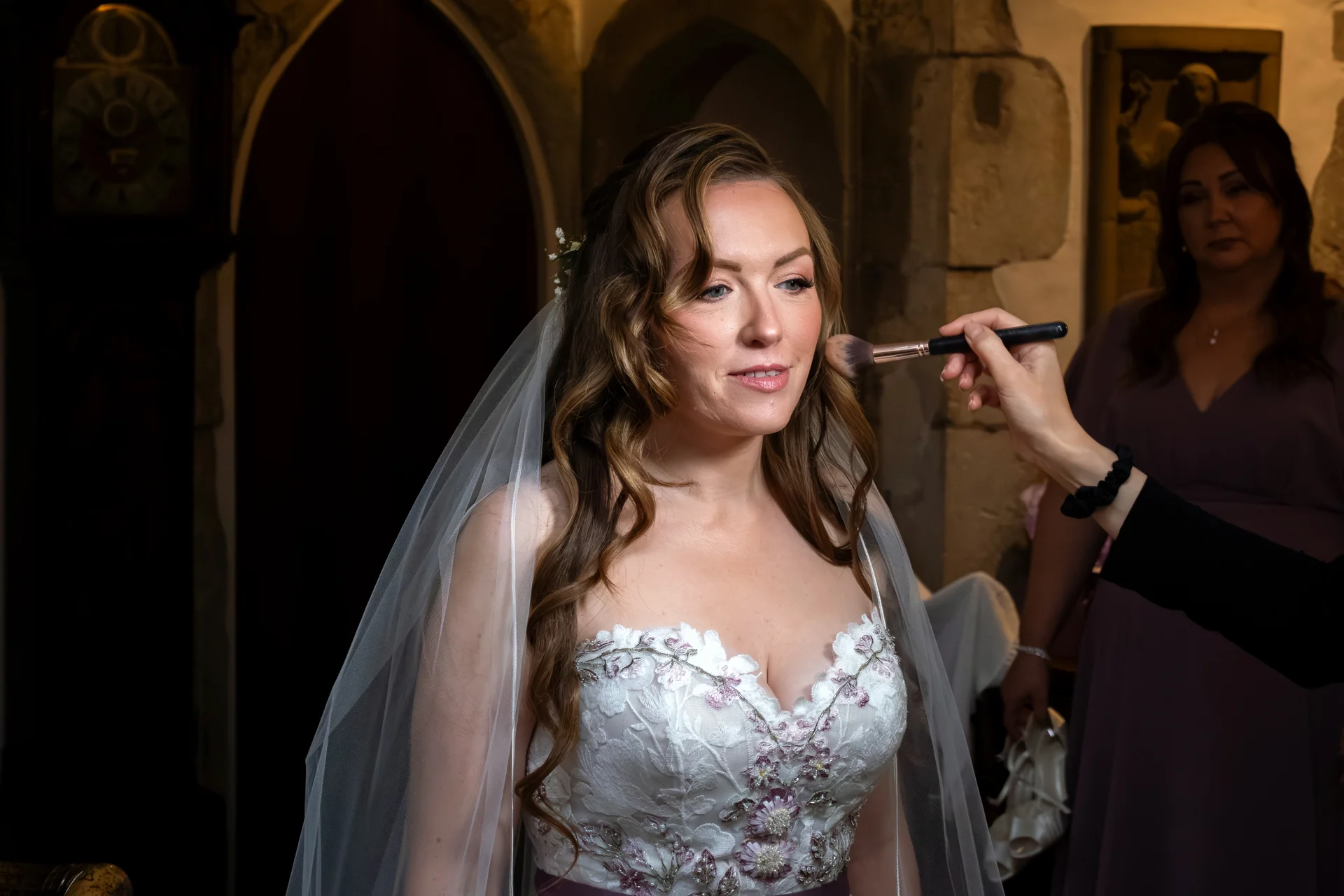 A glowing bride receives her final makeup application during morning preparations, wearing an embroidered floral gown and a delicate veil in a stone interior.