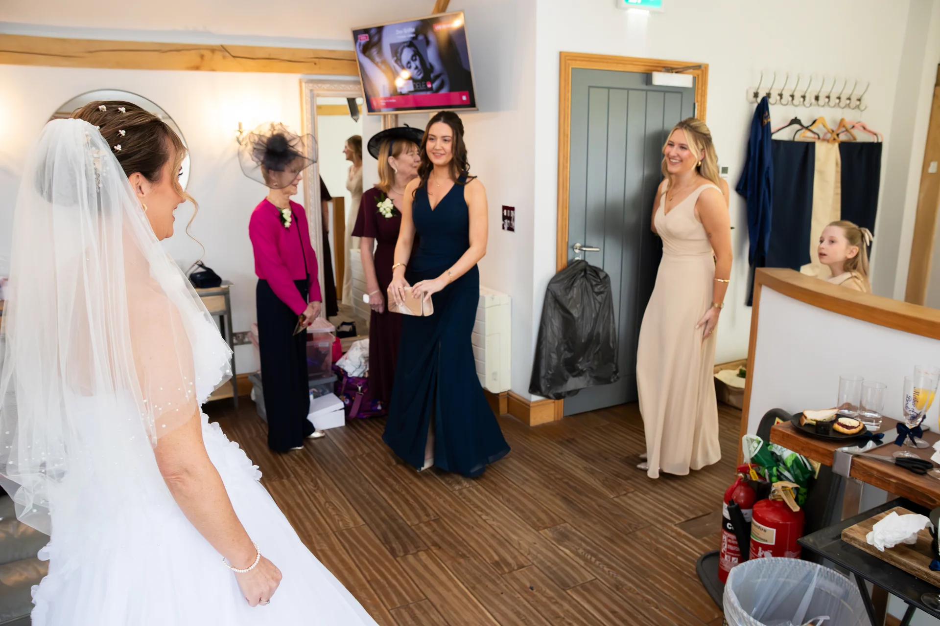 A smiling bride wearing a white gown and veil stands in a room while her bridesmaids, mother, and grandmother look on admiringly during morning preparations.