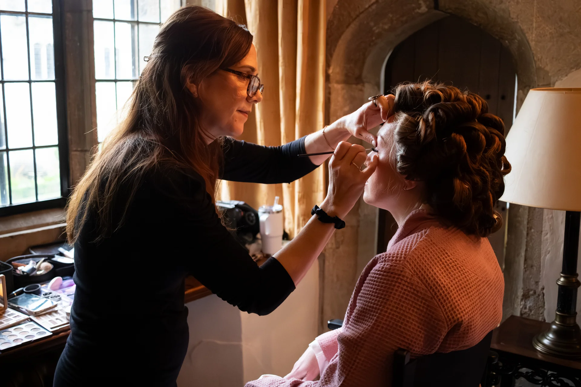 A professional makeup artist carefully applies eyeliner to a bride wearing a pink waffle-textured robe in a historic manor house room with natural light streaming through a grid window.