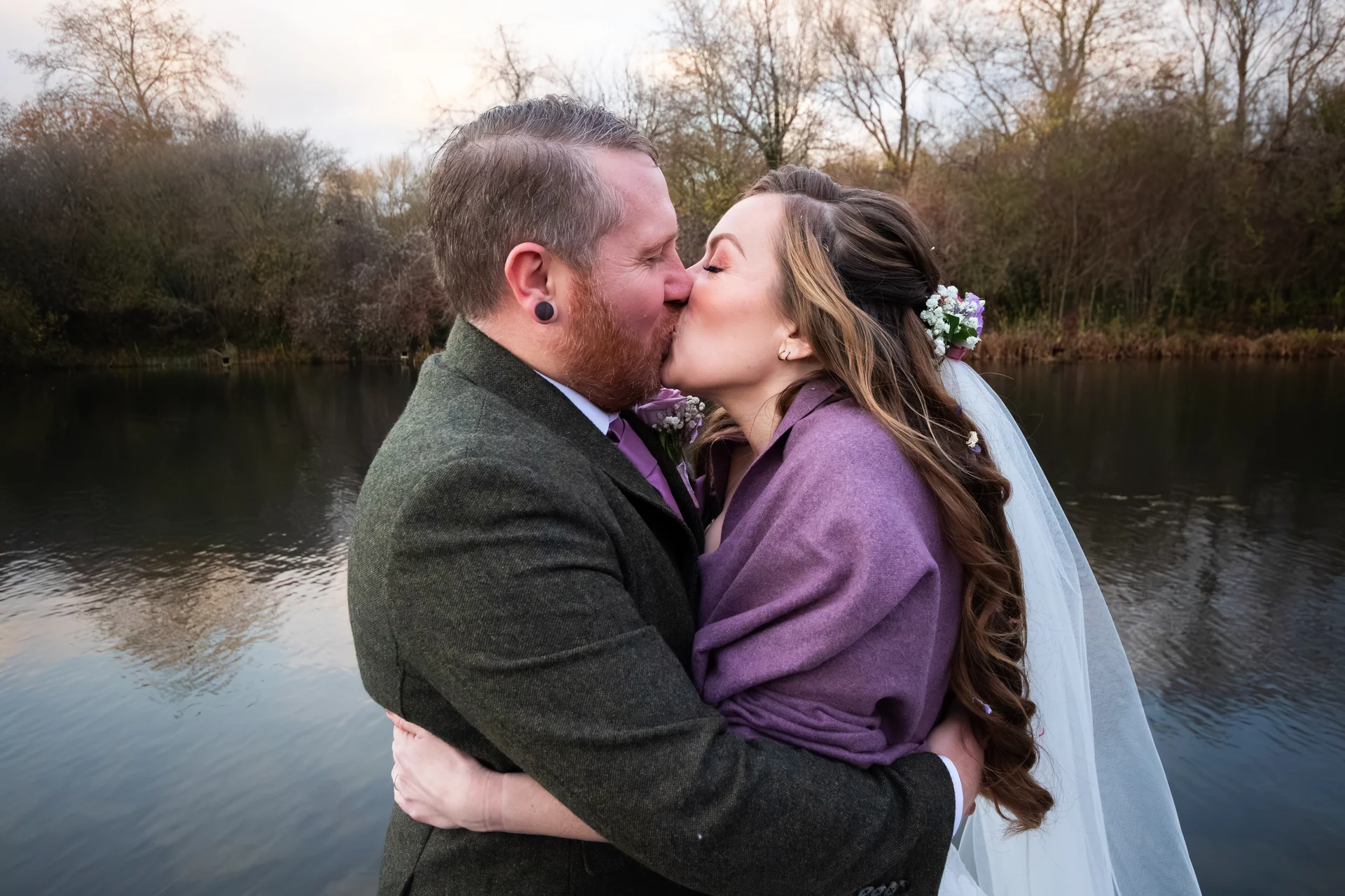 A newlywed couple shares a tender kiss by a tranquil lake during their autumn wedding, with the groom wearing a green tweed suit and the bride wrapped in a purple stole.