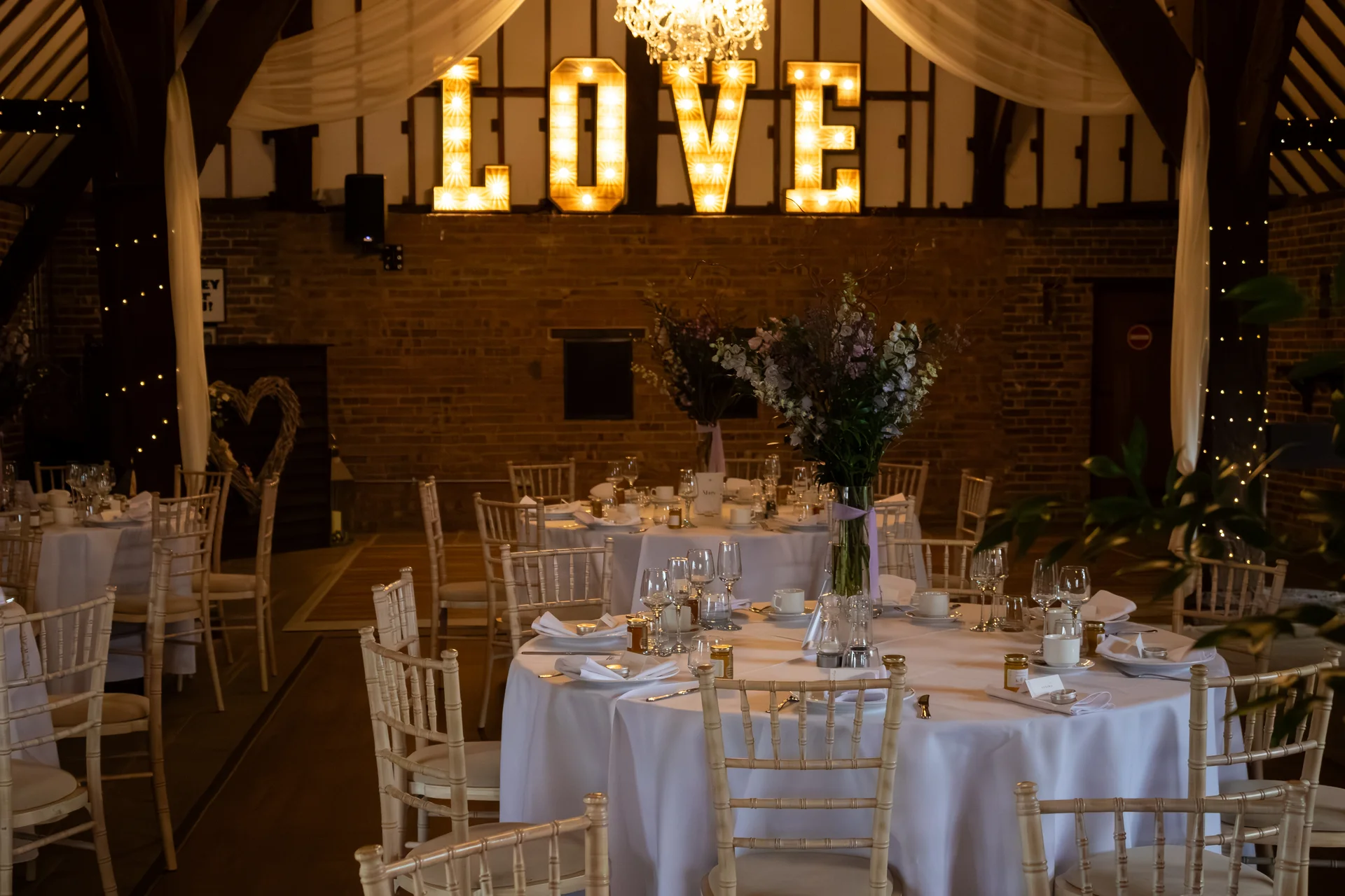 A warmly lit rustic barn wedding reception featuring round tables set with white linens, tall floral centrepieces, and a large illuminated love sign against an exposed brick wall.