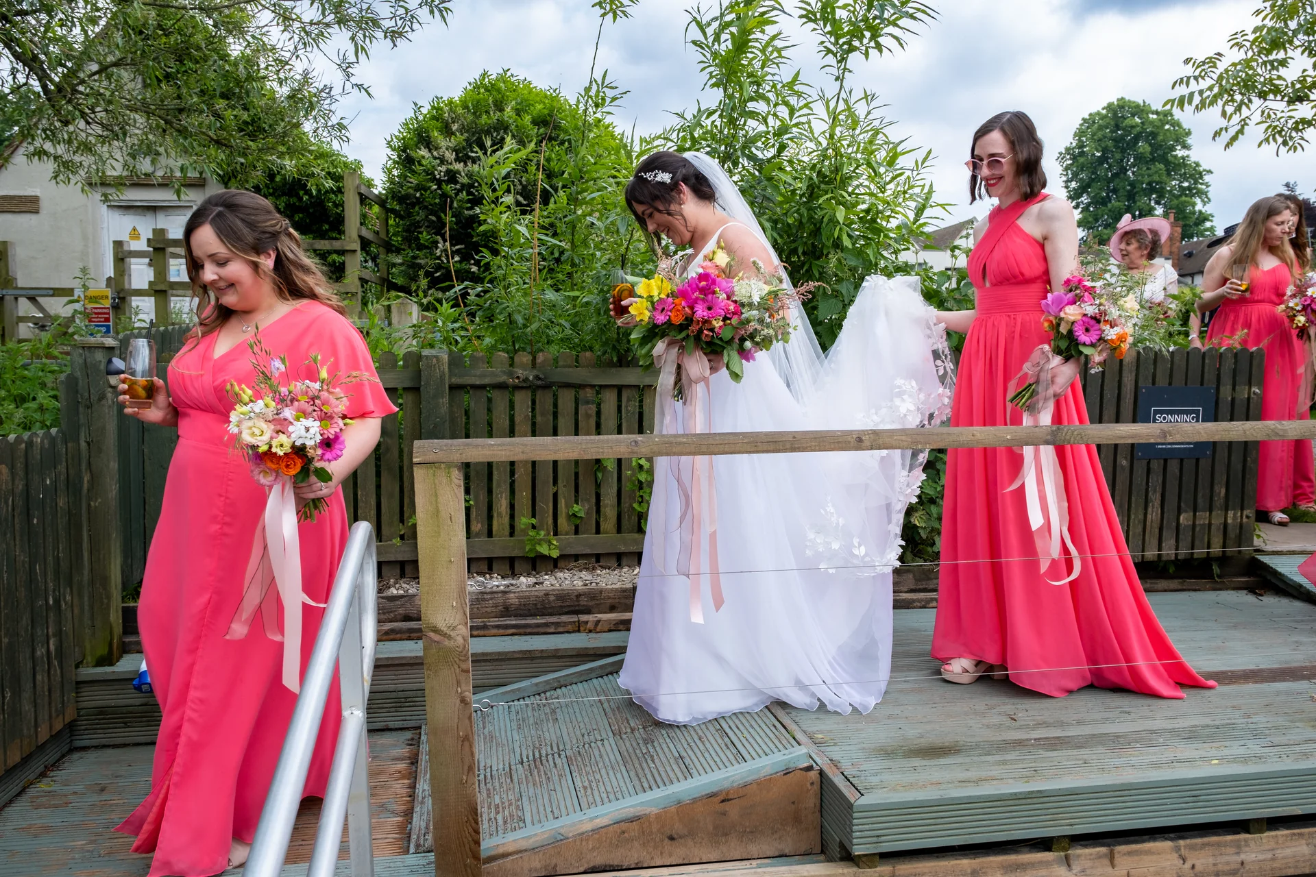 A smiling bride wearing a white gown and veil walks across a wooden footbridge alongside bridesmaids dressed in vibrant pink dresses holding colourful bouquets.