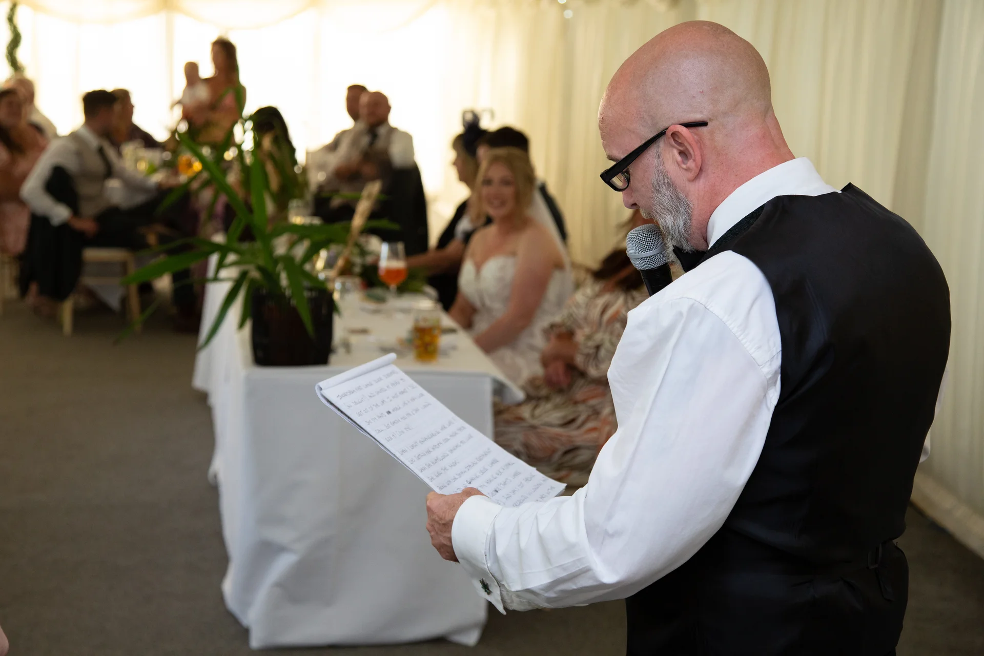 A bald speaker wearing a white shirt and black waistcoat reads a speech from paper notes while holding a microphone at a wedding reception.