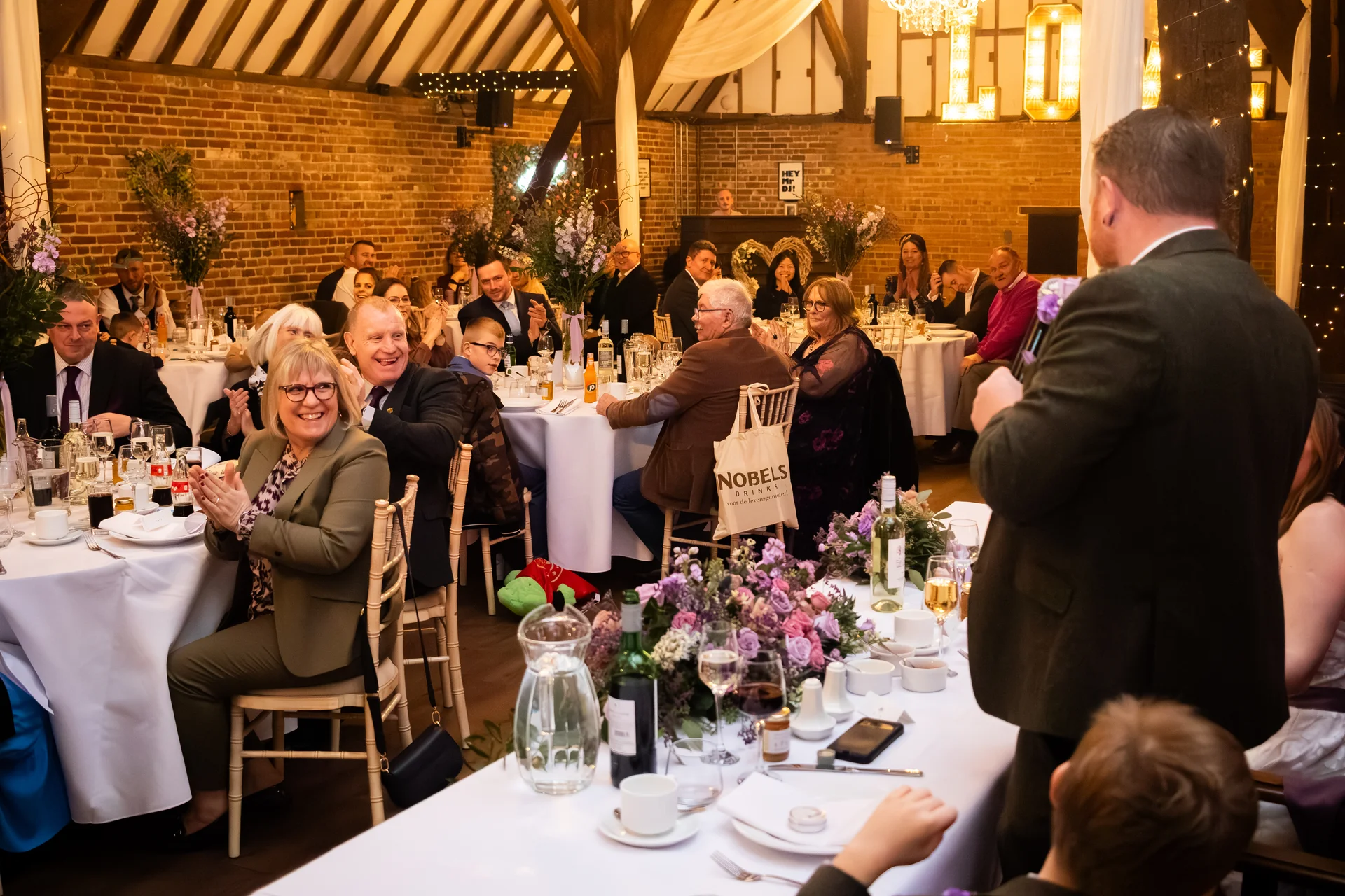 A guest delivers a speech during the wedding reception in a rustic barn with exposed brick and wooden beams, while seated guests laugh and applaud around banquet tables.