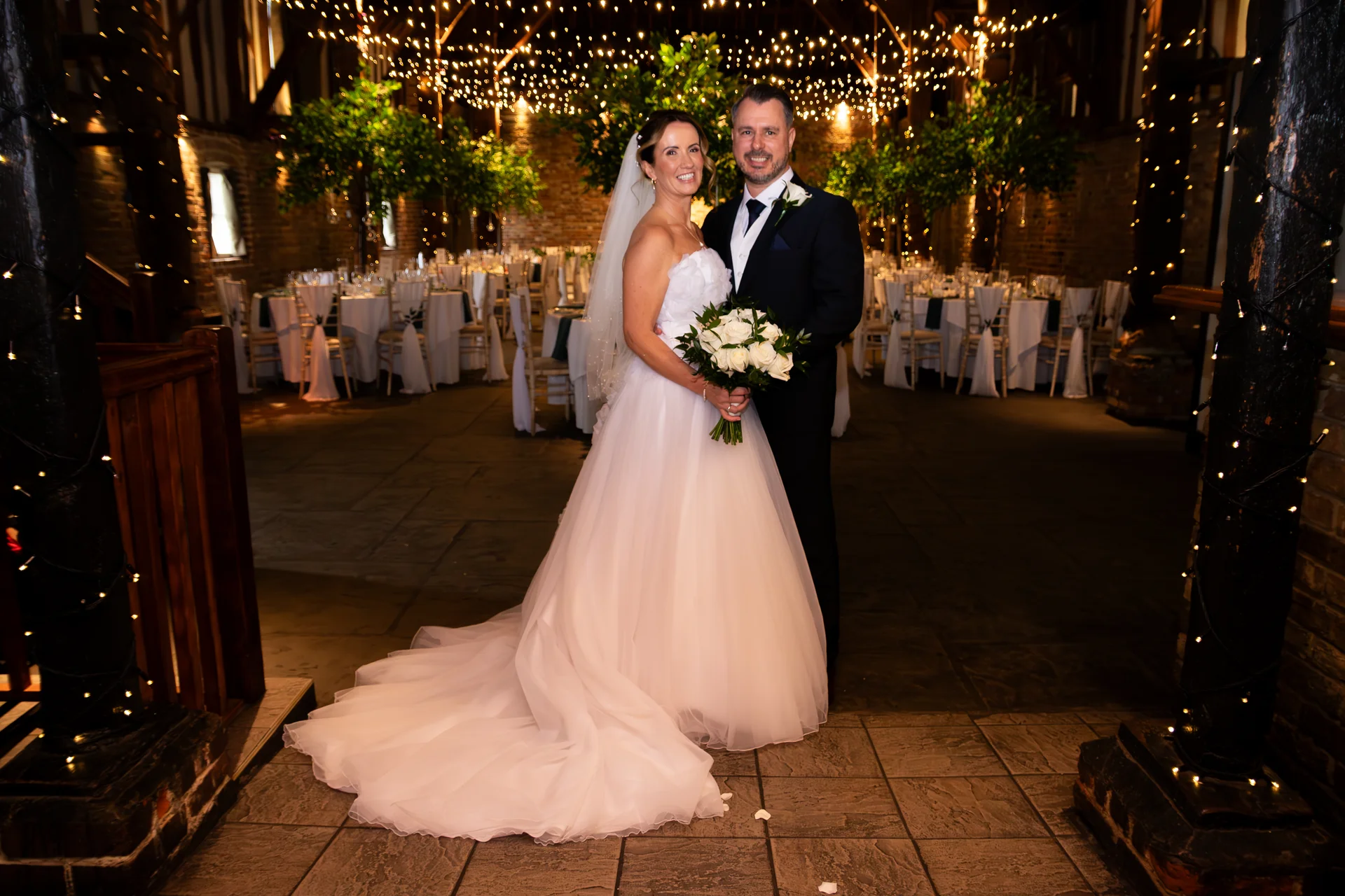 A newlywed couple poses inside a rustic barn wedding venue illuminated by fairy lights, with the bride holding a bouquet of white roses.