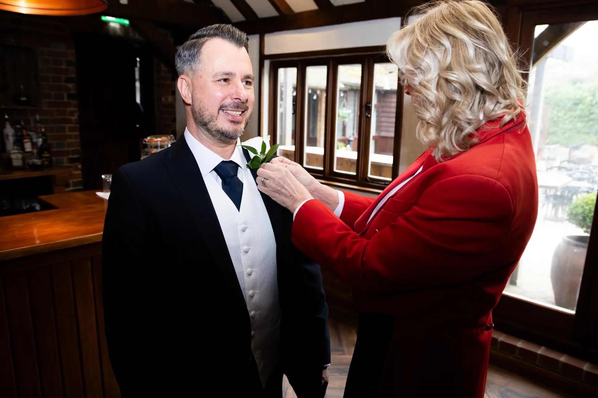 A groom in a black suit and white waistcoat smiles as a woman in a red jacket pins a white boutonniere to his lapel indoors.