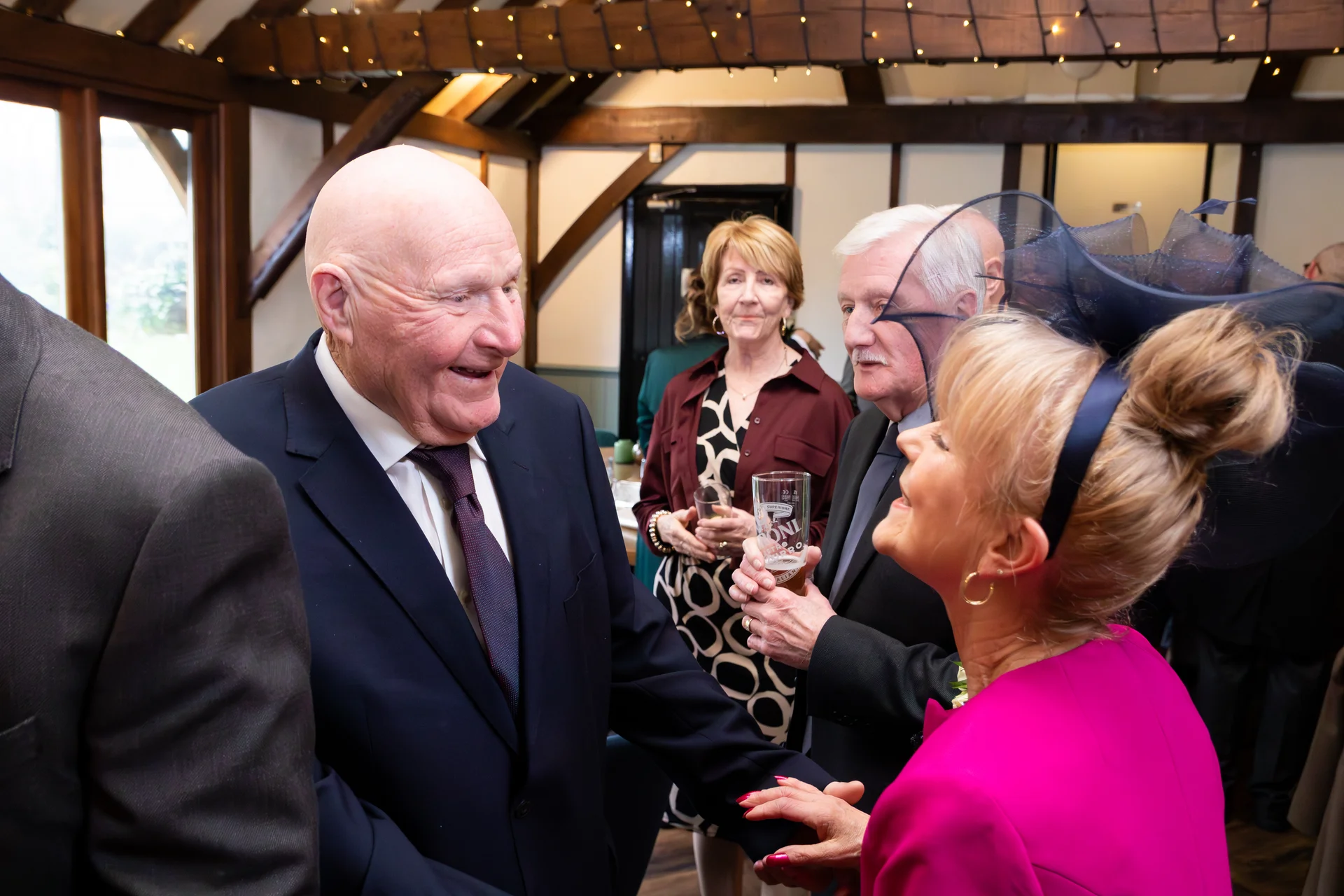 Older wedding guests smiling and mingling inside a rustic barn venue with exposed timber beams and fairy lights.