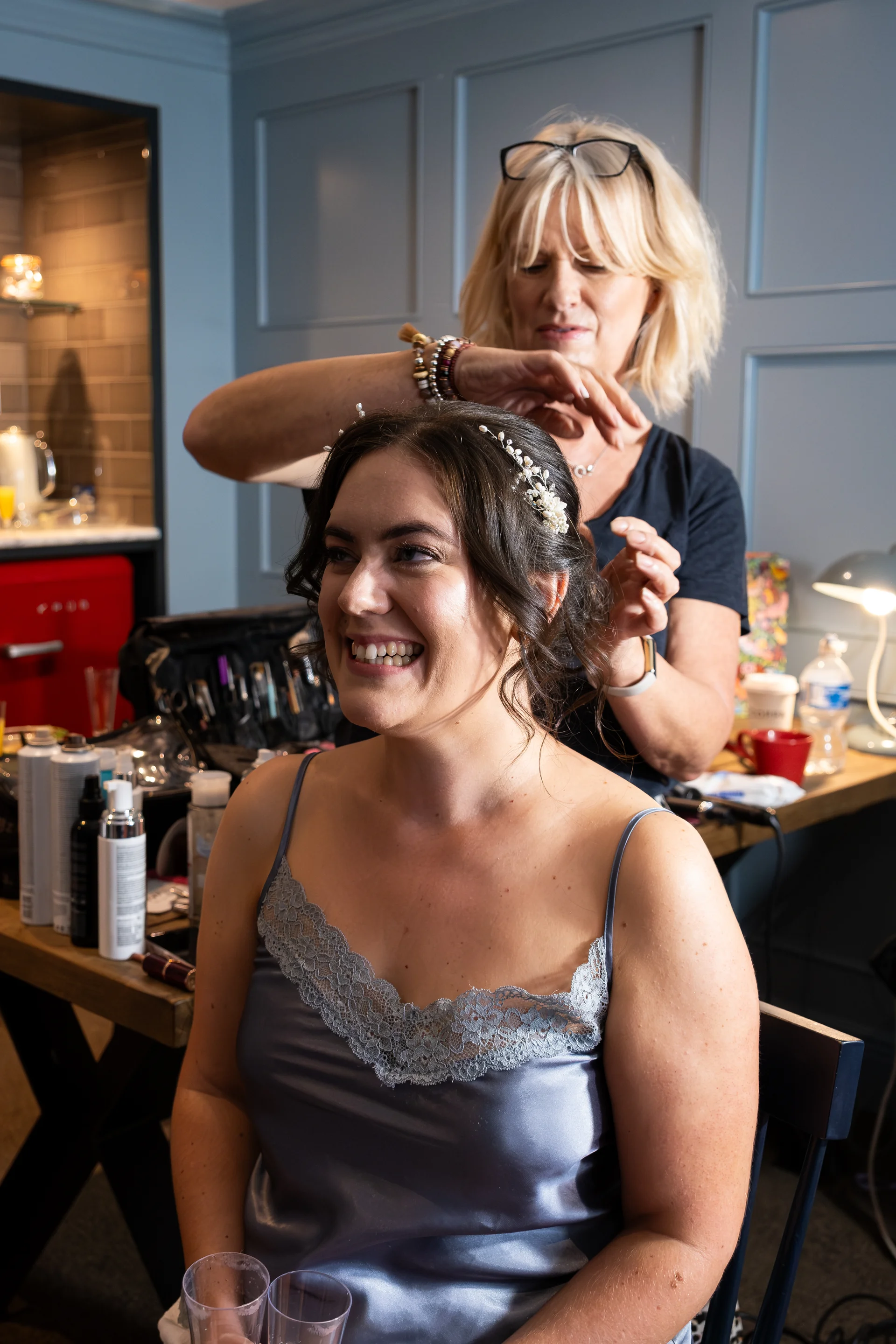 A smiling bride with a delicate pearl hair vine enjoys getting ready in a bridal suite while a stylist expertly arranges her hair.