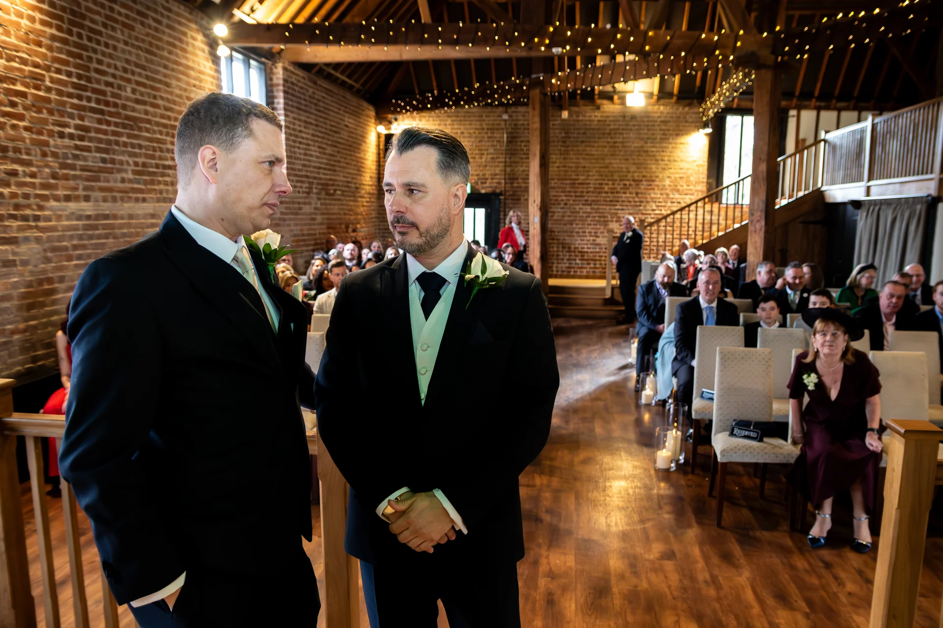 Two grooms stand at the altar inside a rustic oak-framed barn with exposed brick walls and a fairy light canopy, looking at each other during their wedding ceremony while guests watch from seated rows.