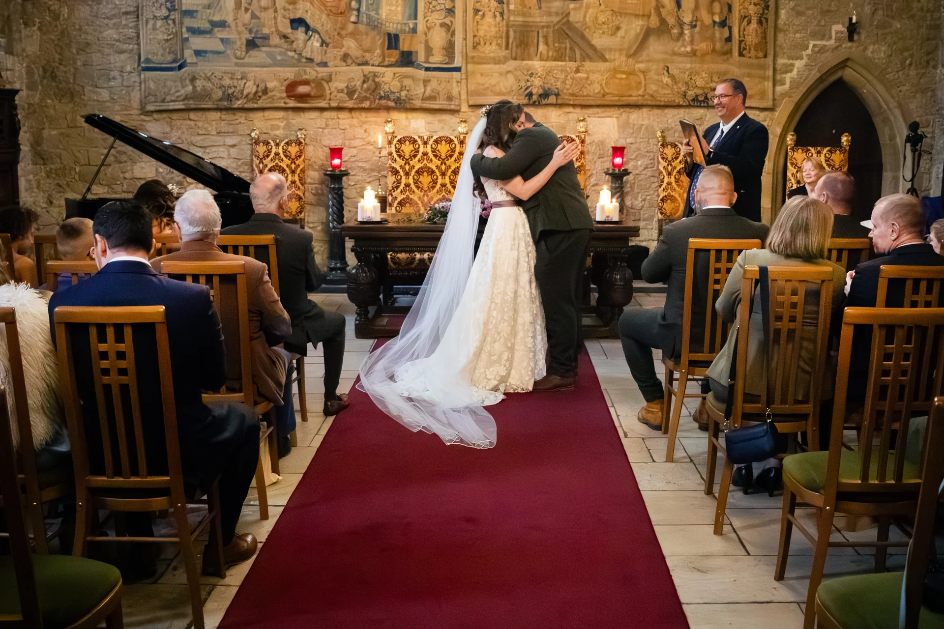 A bride and groom embrace on a red carpet in a historic stone castle ceremony room while guests watch from wooden chairs.