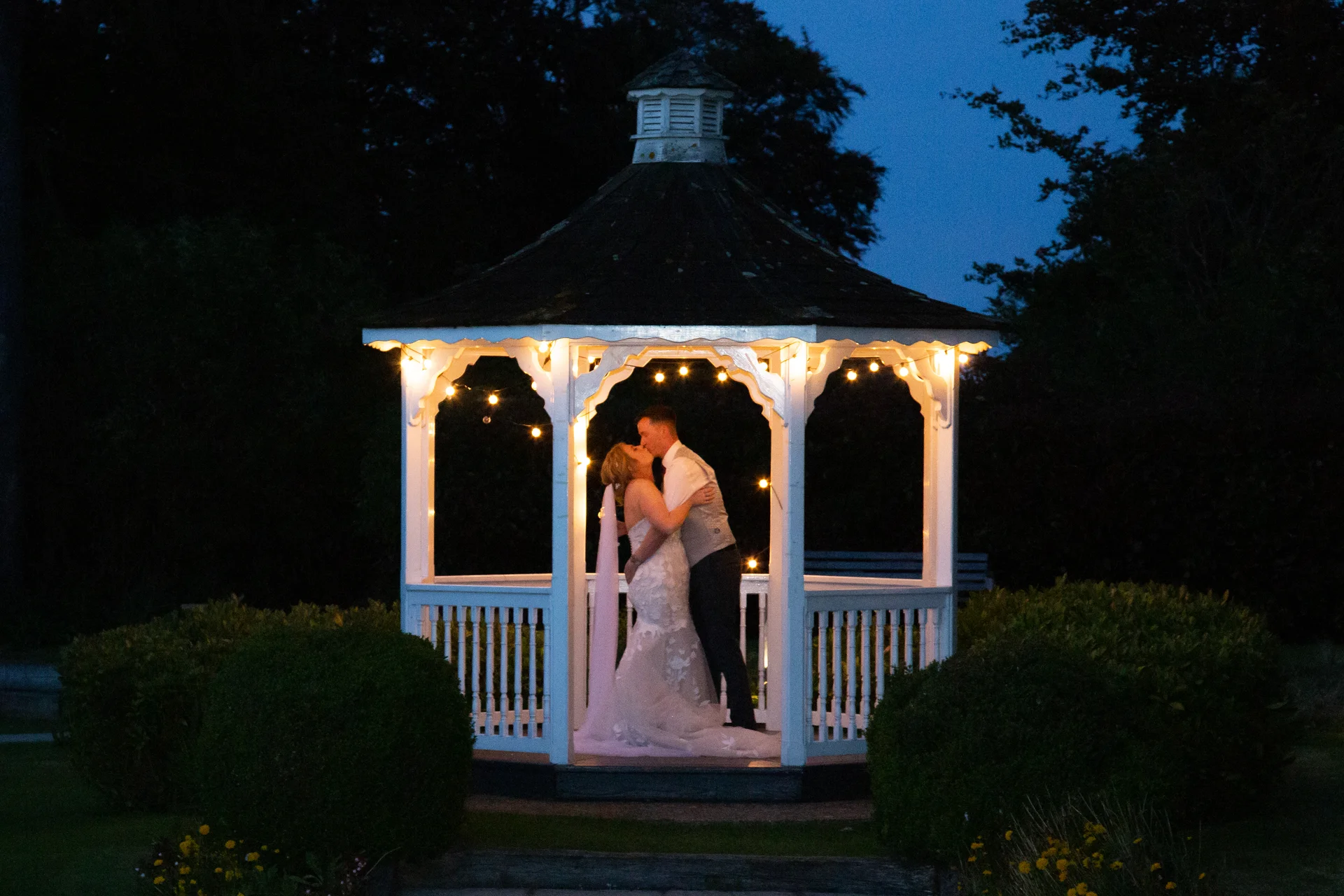 A newlywed couple shares a romantic kiss inside an illuminated white gazebo during twilight blue hour.