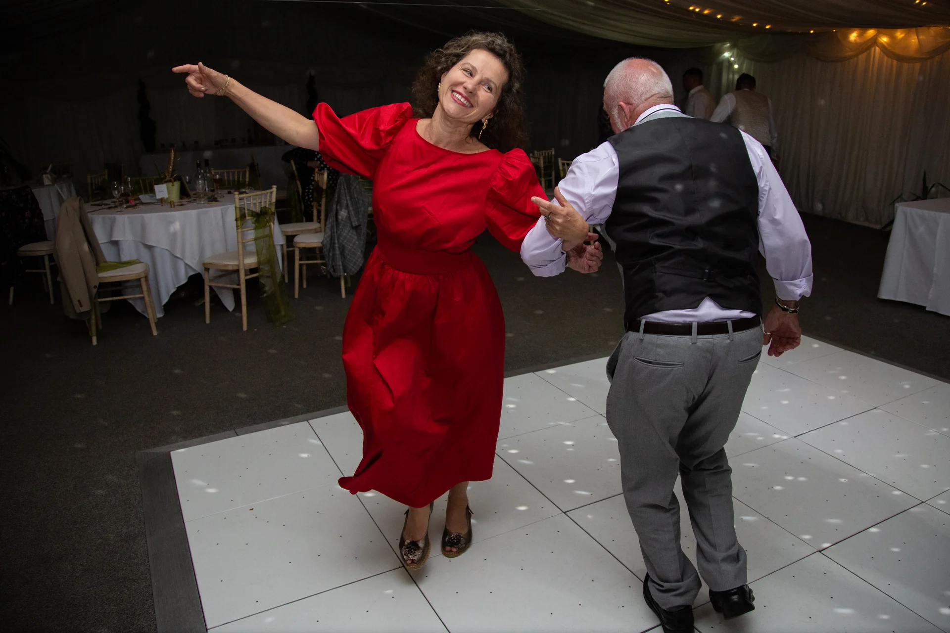 An older couple dancing energetically together on a glowing white dance floor inside a marquee reception.