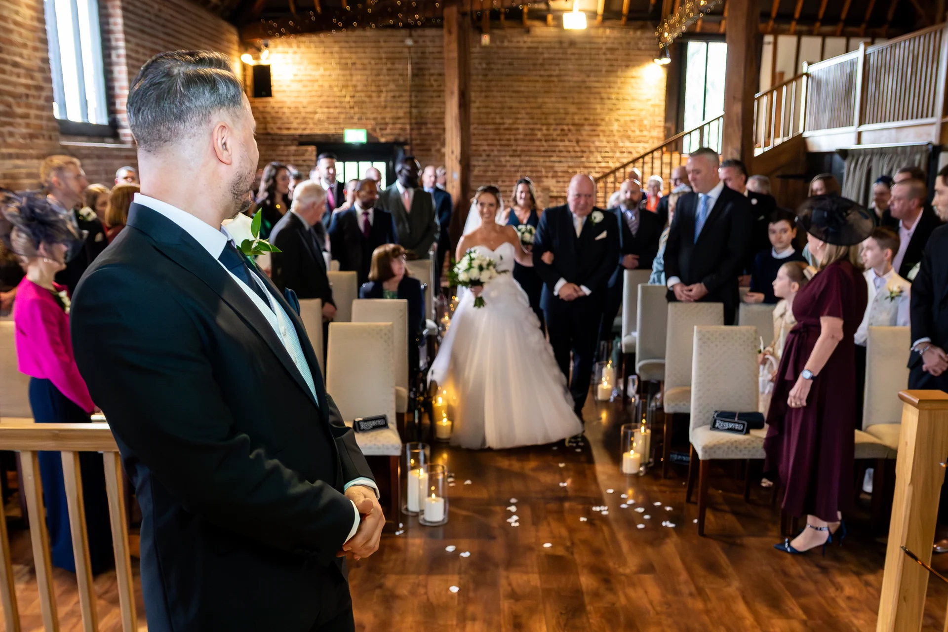The groom looks back from the altar as the bride walks down the aisle accompanied by her father inside a rustic converted barn wedding venue.