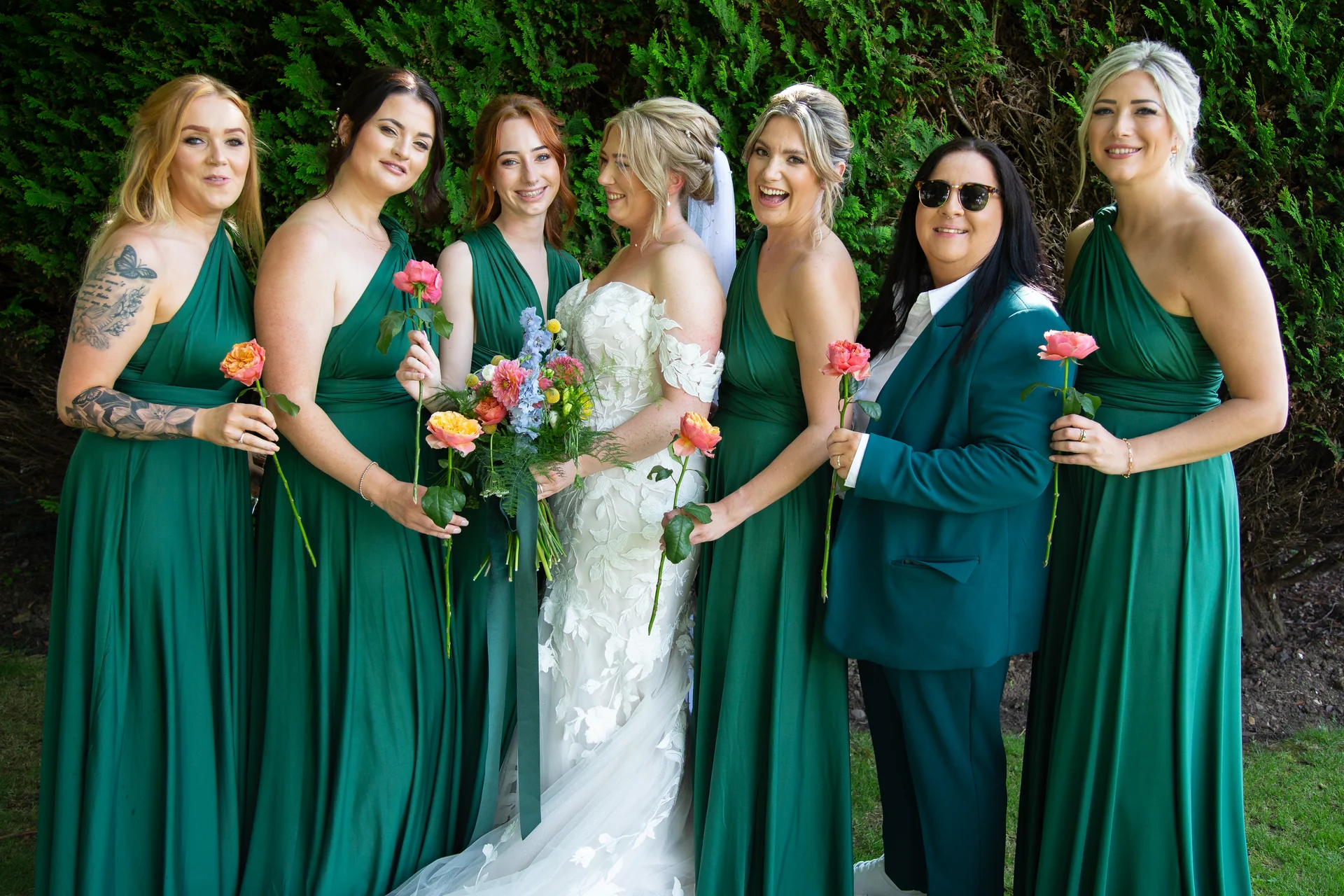 A joyful bride in a white lace gown stands flanked by bridesmaids in matching emerald green dresses and a groomsman in a green suit outdoors against a lush green hedge.