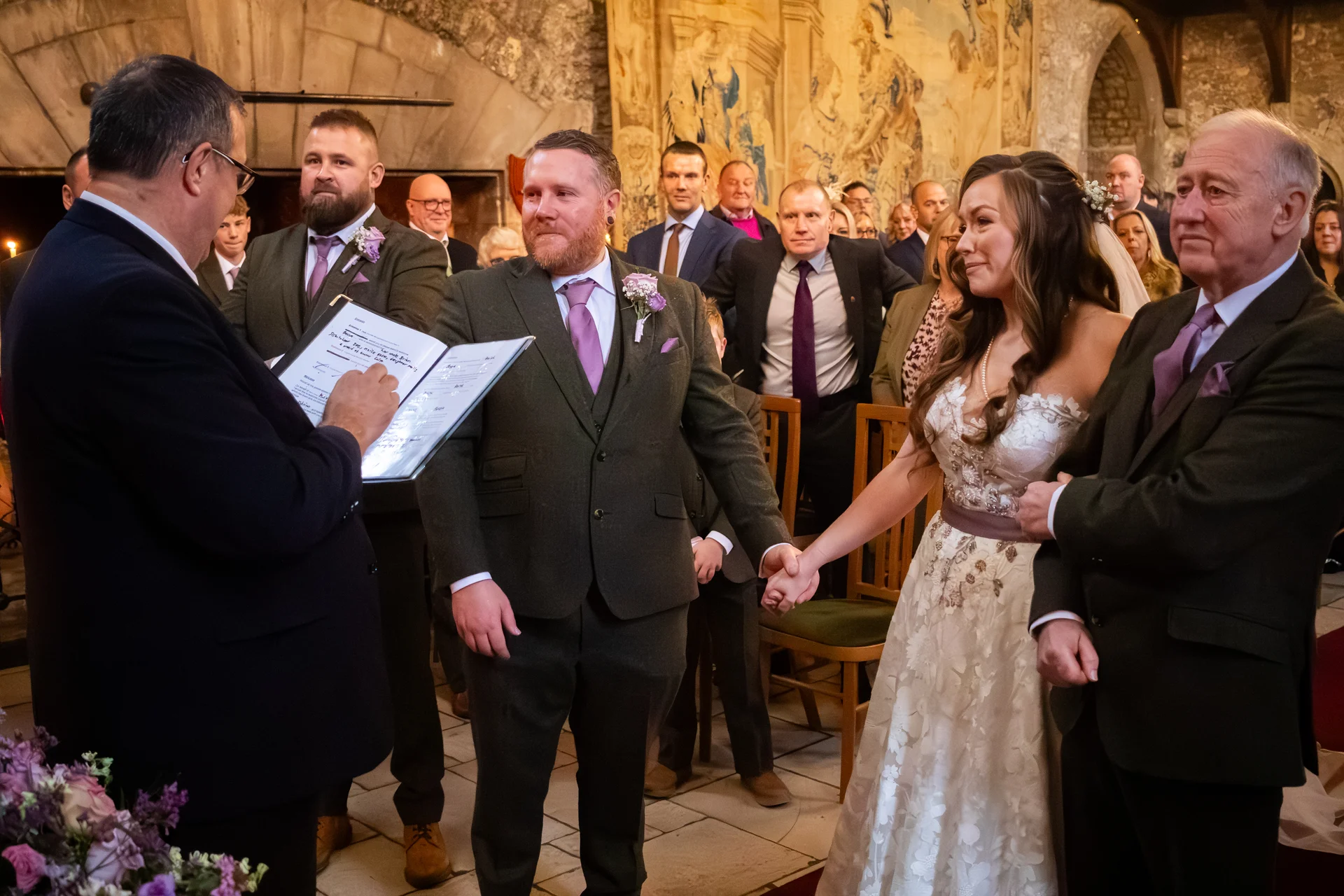 The bride and groom hold hands during the signing of the register at their historic venue ceremony, surrounded by family and wedding party members.