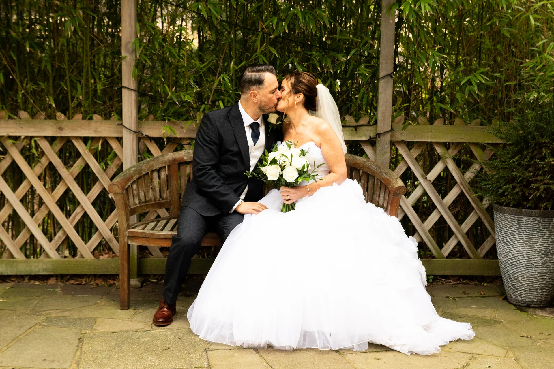 A bride and groom share a tender kiss while seated together on a wooden garden bench outdoors.
