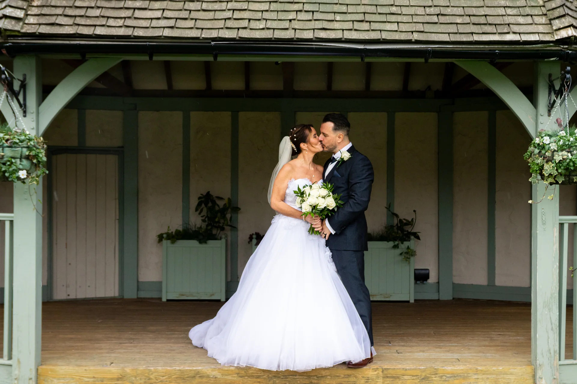 Newlywed bride and groom share a tender kiss underneath a green-framed wooden gazebo holding a white rose bouquet.