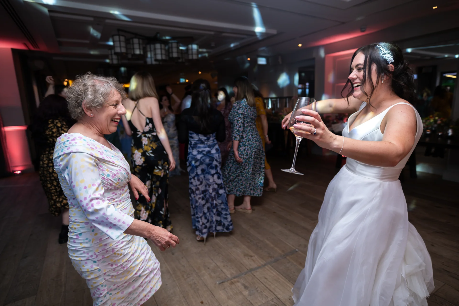 A joyful bride dances energetically on the dance floor holding a glass of wine, laughing alongside an older guest at the evening wedding reception.