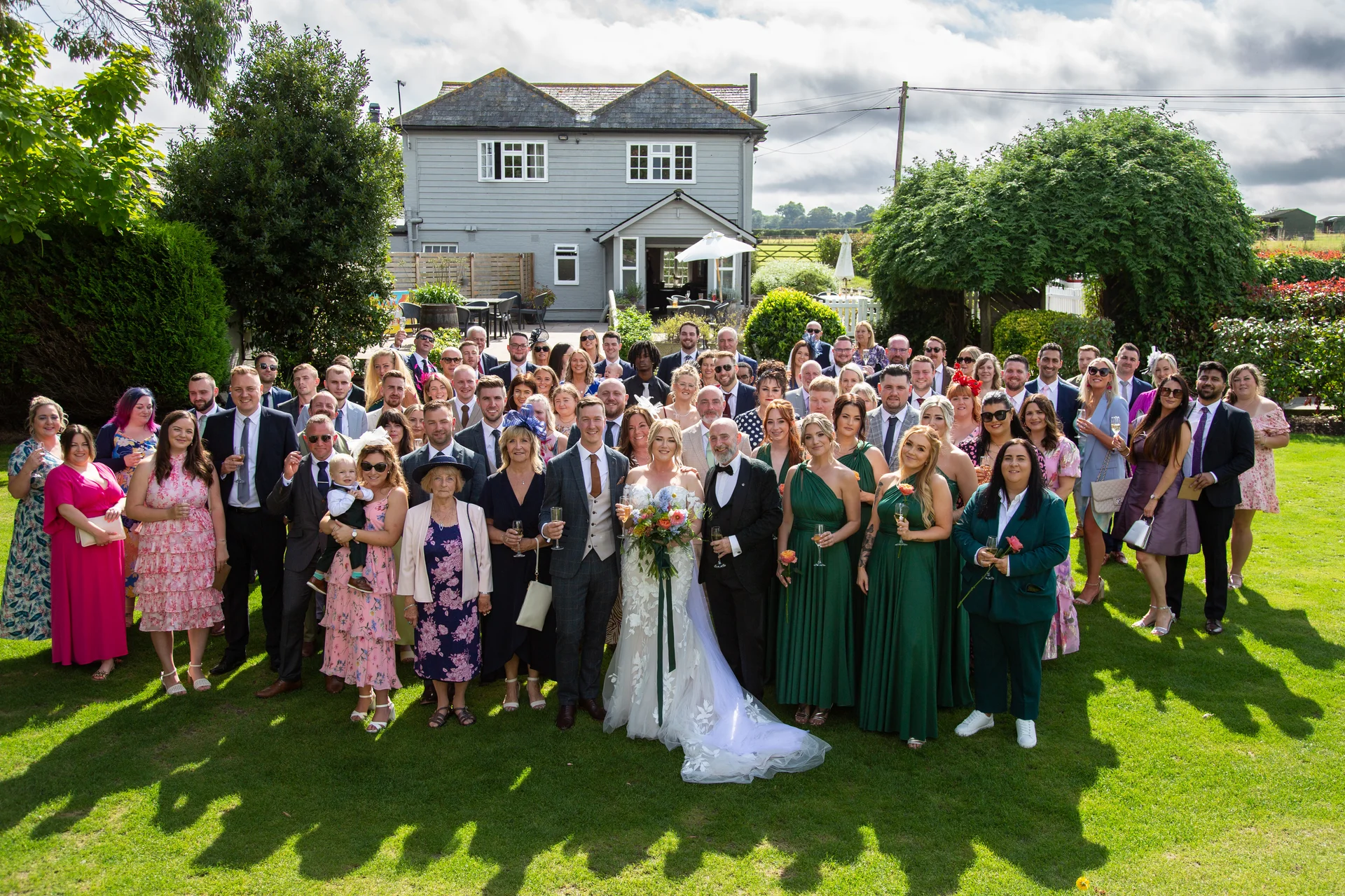 A large wedding party and group of guests pose together on a manicured lawn in front of a country house on a sunny day.
