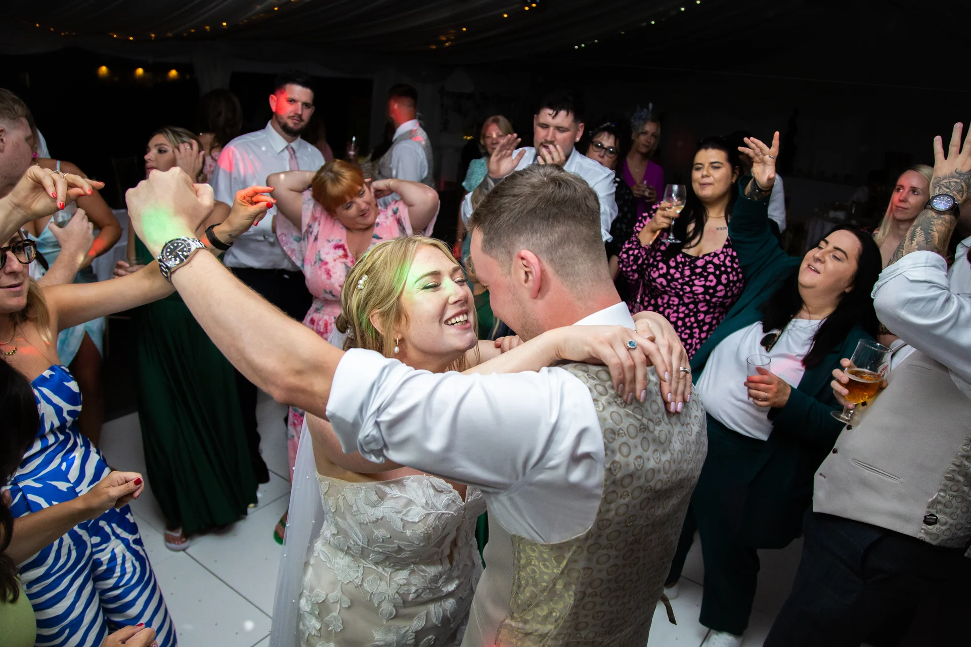 A newlywed couple laugh and dance joyfully together on a packed dance floor surrounded by celebrating wedding guests under party lighting.