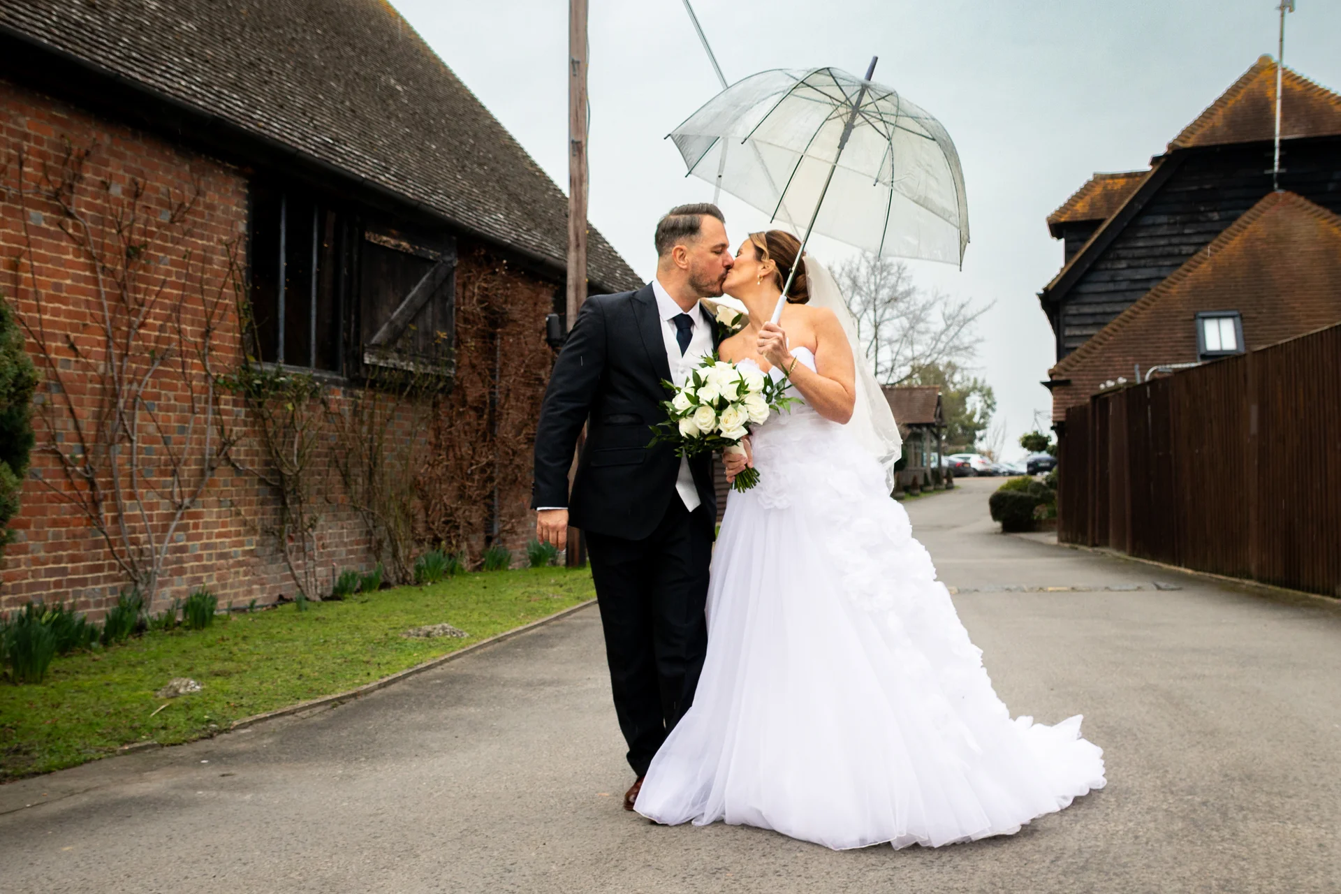Newlyweds share a romantic kiss under a clear umbrella while walking along a path next to a rustic brick barn on their rainy wedding day.