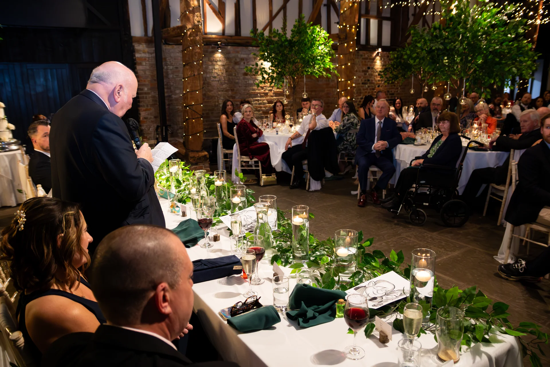 An older gentleman delivers a heartfelt speech during the wedding reception inside a rustic barn decorated with exposed brick and twinkling fairy lights.
