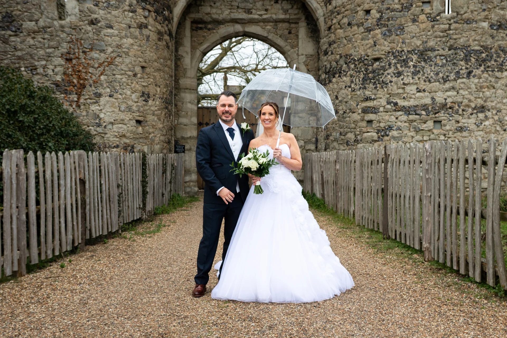 A newlywed couple smiles together under a clear umbrella on a gravel path in front of a historic castle archway and stone walls.