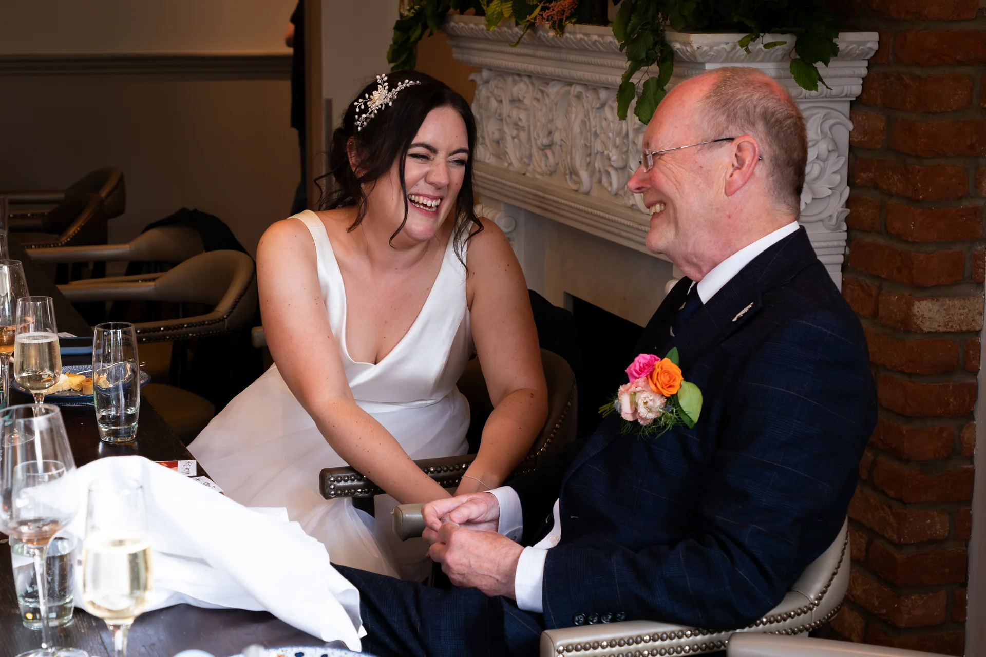 A joyful bride in a white gown and a smiling guest in a dark patterned suit share a hearty laugh seated together at the wedding reception.