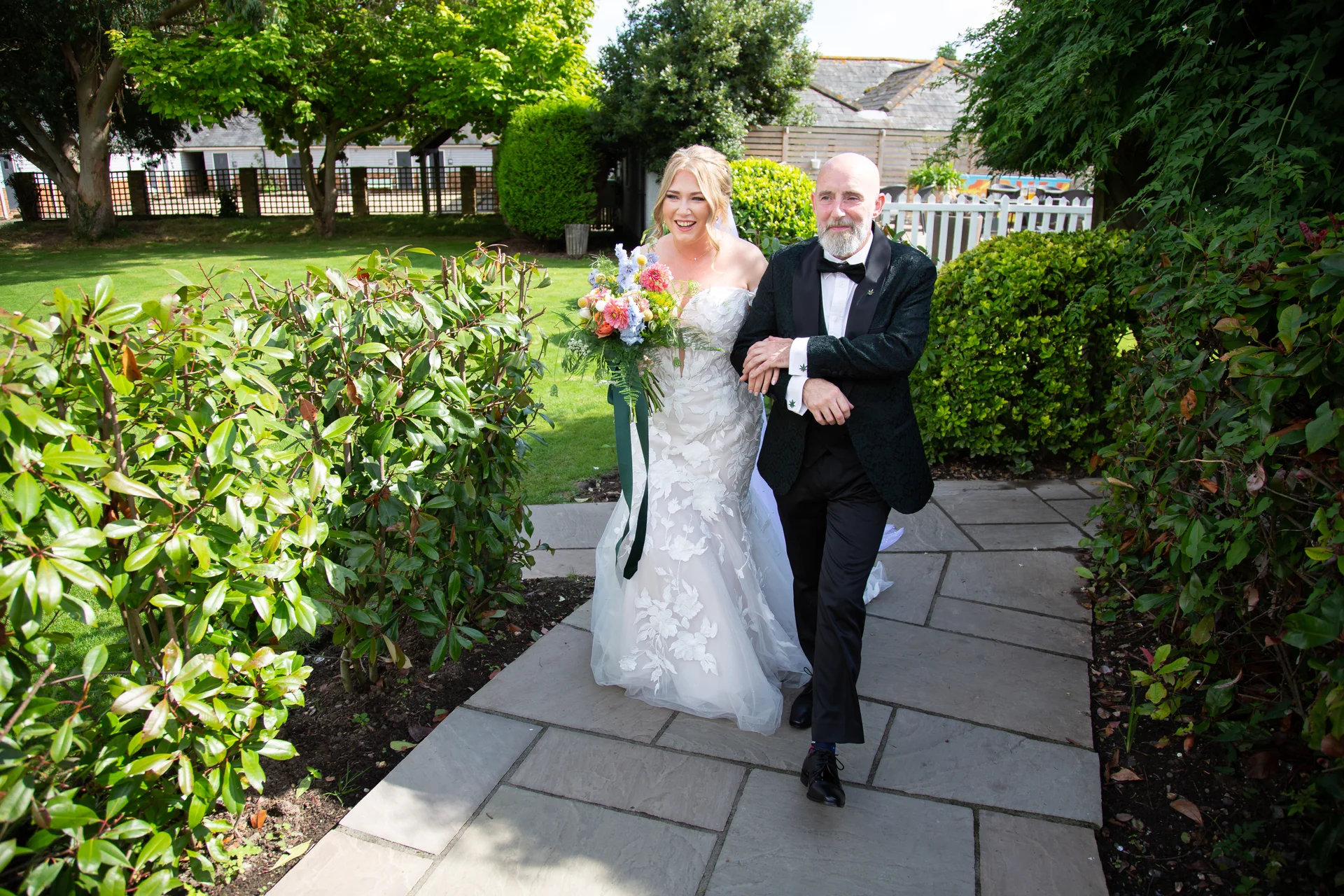 A smiling bride holding a colourful bouquet walks arm in arm with her father along a stone pathway in a lush garden.