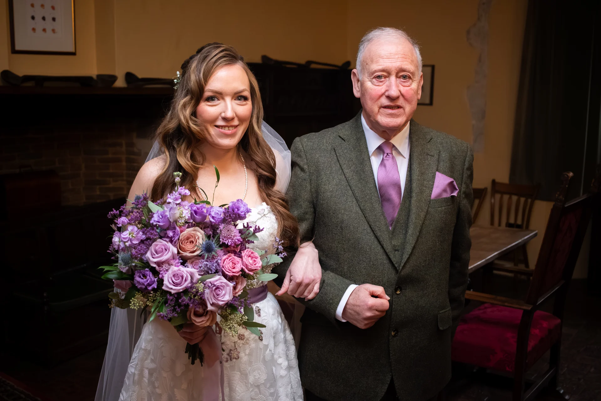 A smiling bride holding a purple floral bouquet links arms with her father, who is wearing a green tweed suit and purple tie.