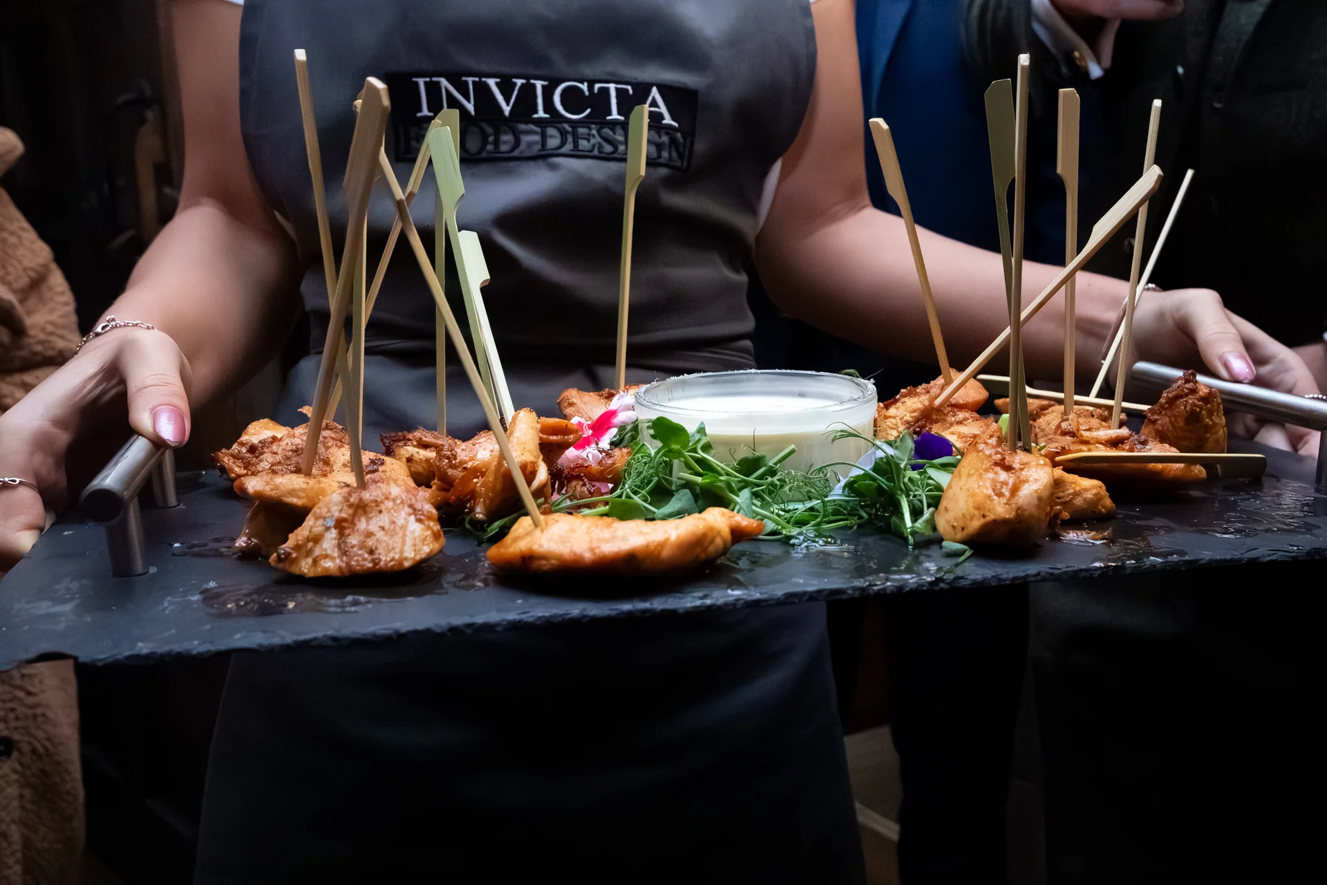 A member of catering staff wearing an Invicta Food Design apron carries a slate platter loaded with skewered chicken canapes and a central dipping sauce during a wedding reception.