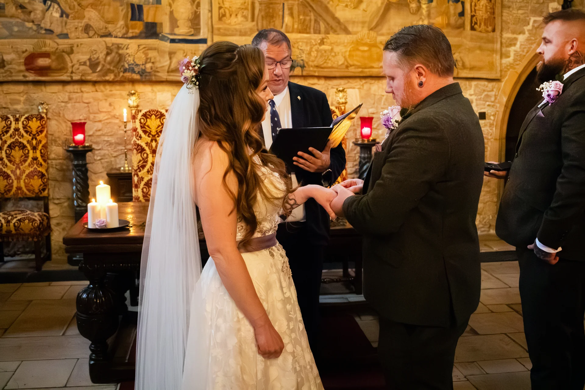 The groom places a ring on the bride's finger during a candlelit ceremony inside a historic stone hall.