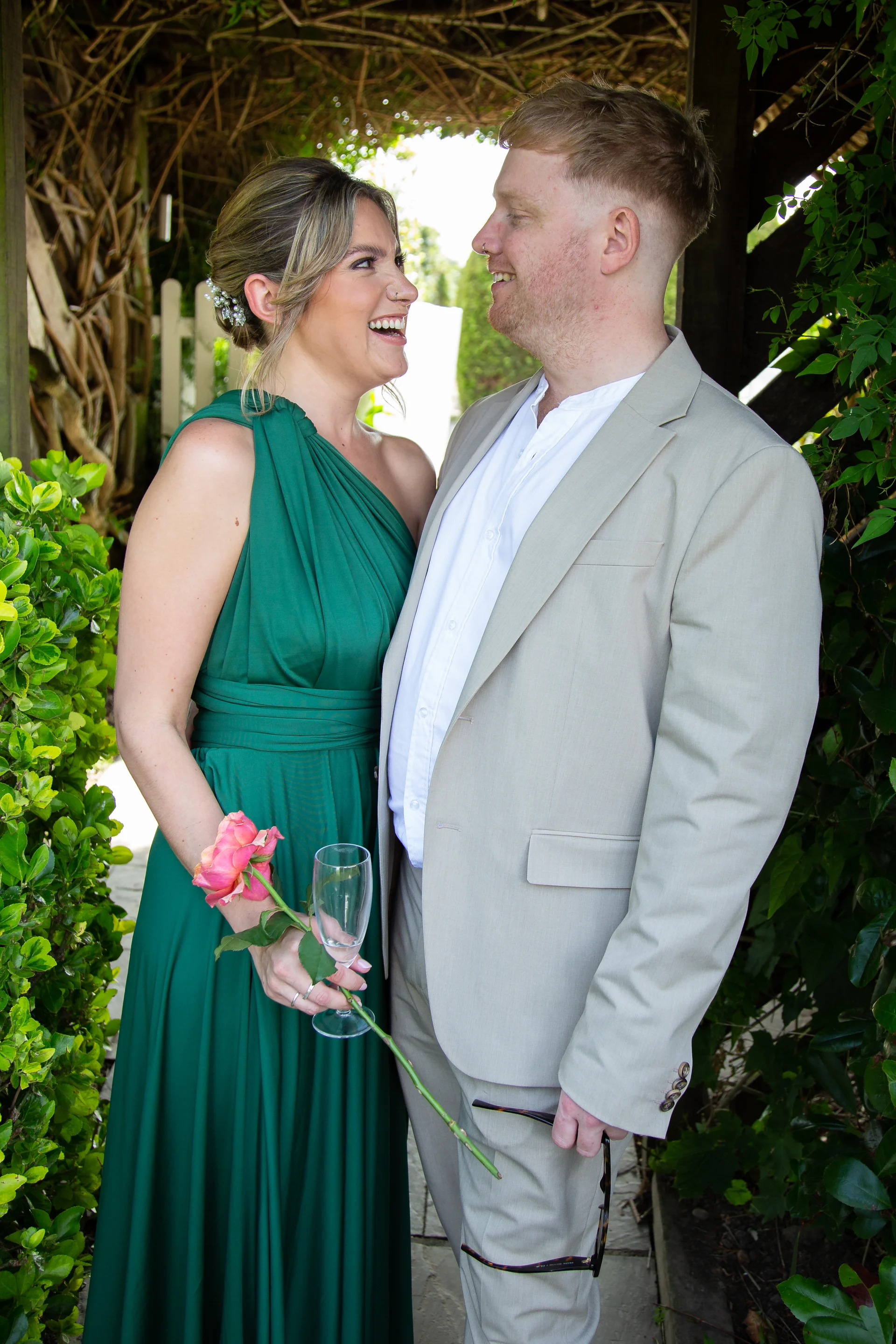 A smiling couple shares a joyful moment outdoors, with the bride wearing a vibrant green one-shoulder dress and holding a single rose and champagne glass, while the groom wears a stone linen suit.