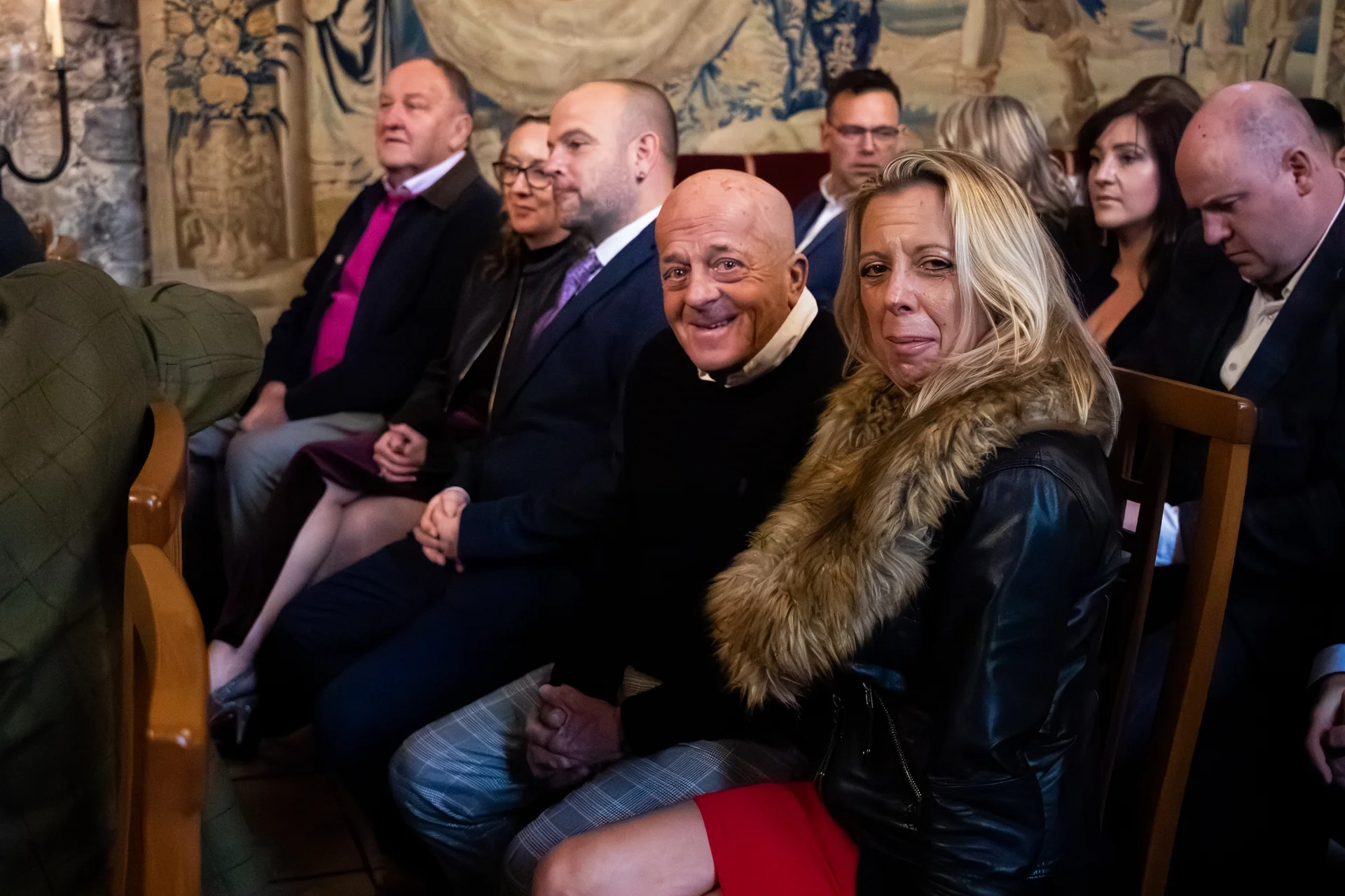 Wedding guests seated closely together in a historic venue watching proceedings, with an older couple smiling towards the camera in the foreground.
