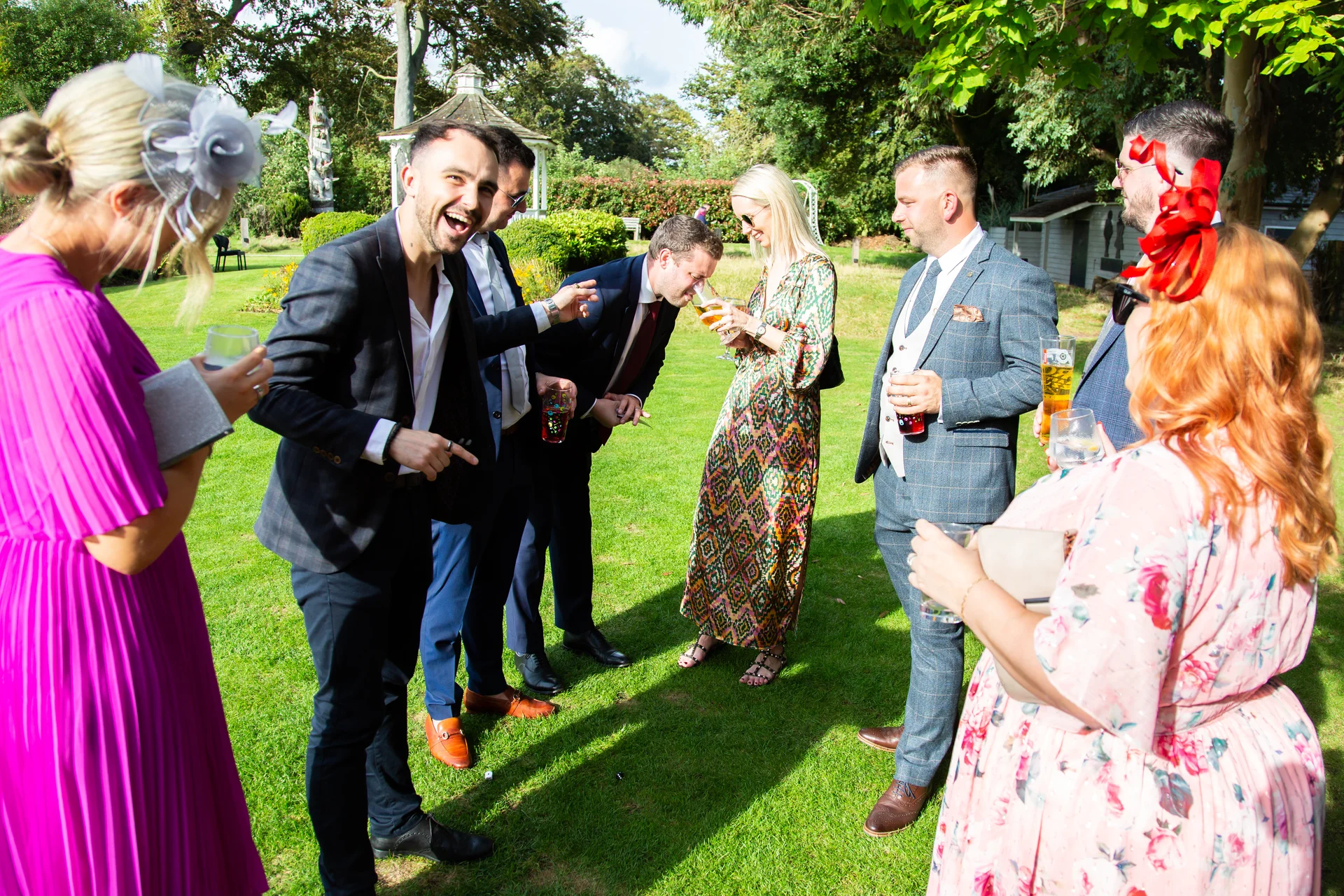 A group of smartly dressed wedding guests laughing and socialising on a sunlit lawn during an outdoor drinks reception.