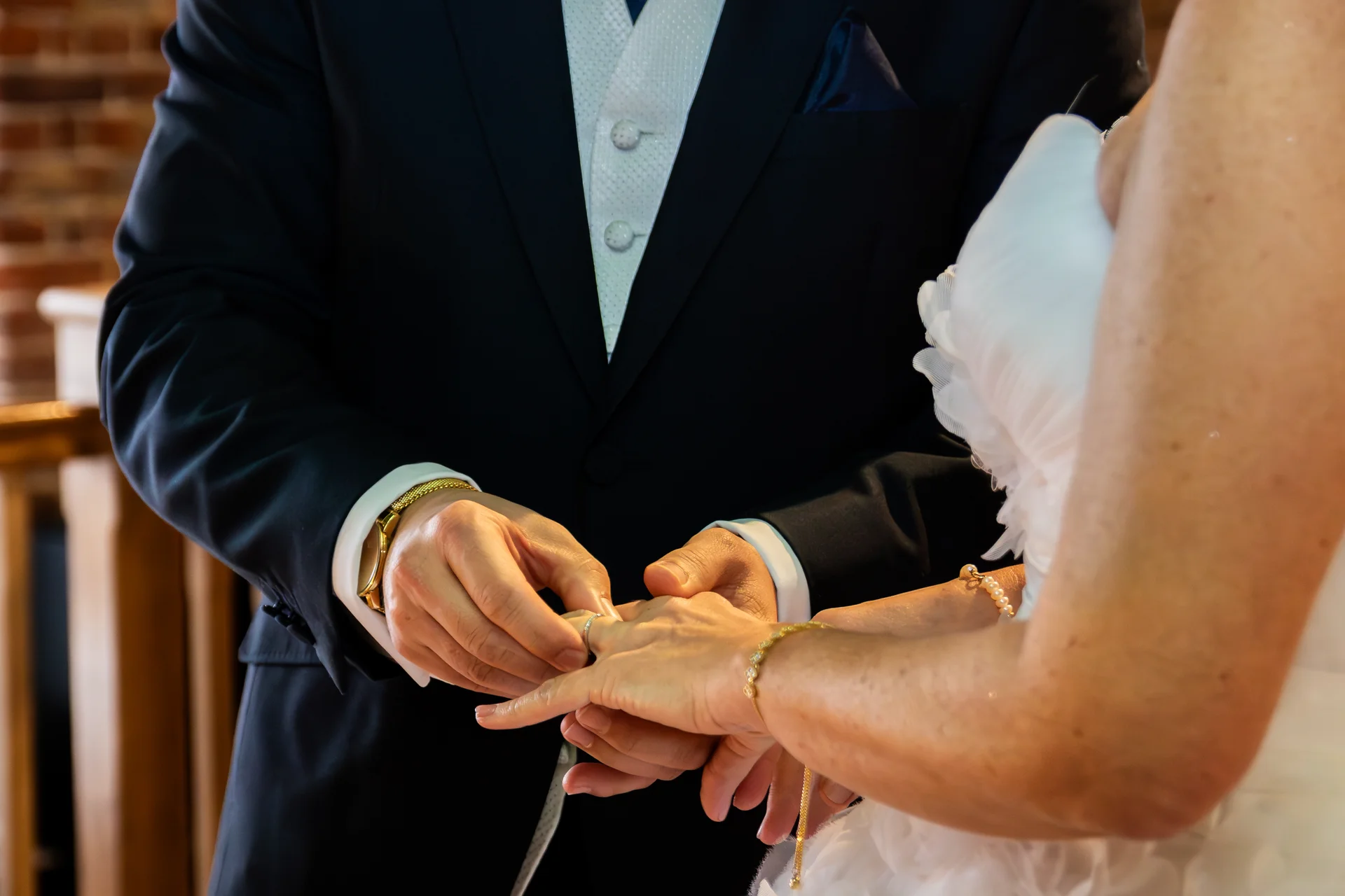 Close-up of a groom in a dark suit and white waistcoat gently placing a wedding ring onto the bride's finger during the ceremony.