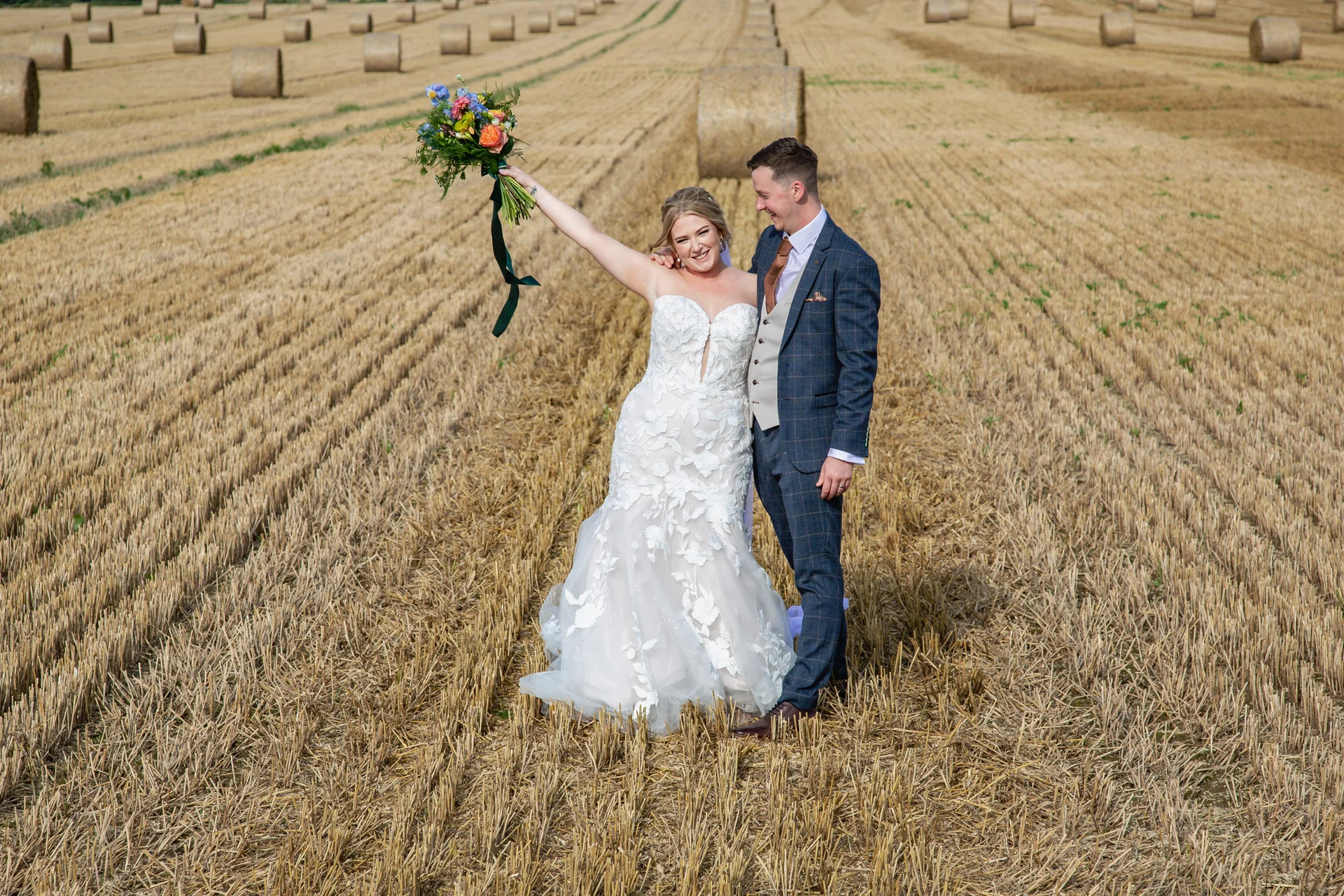 A newlywed couple celebrates standing in a harvested golden field dotted with hay bales, with the bride raising her colourful bouquet in the air.