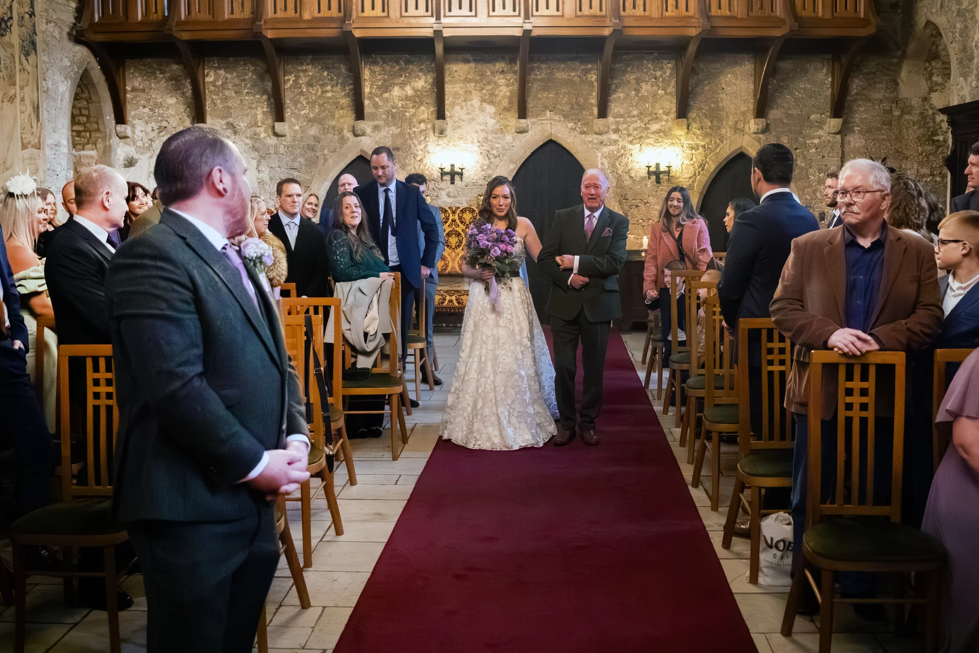 A bride walking down the red carpeted aisle of a historic stone castle venue with her father, watched by seated guests.
