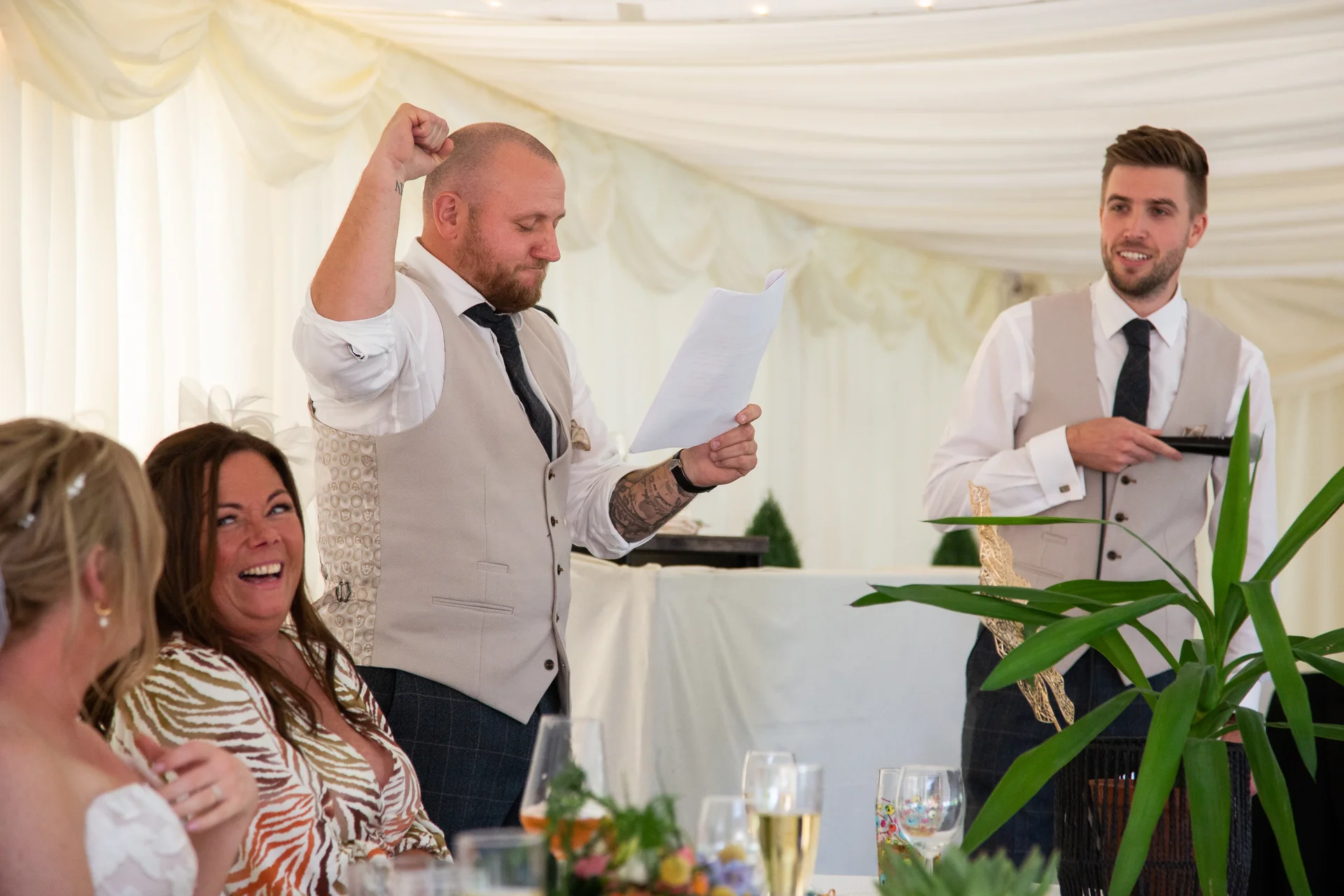 A groom in a tweed waistcoat joyfully raises his fist while reading his speech, as guests laugh and react during the wedding reception inside a draped marquee.