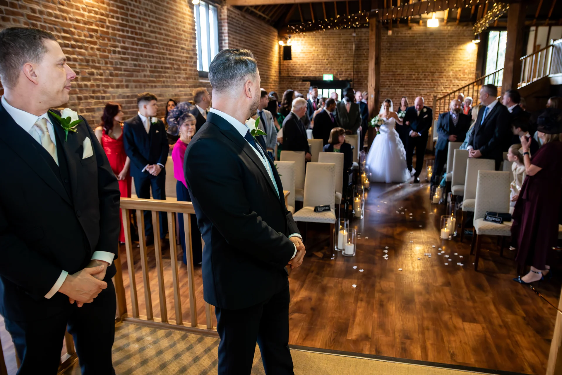 A groom in a black tuxedo looks back as the bride in a classic white gown walks down the aisle of a rustic barn venue decorated with lanterns and exposed brick walls.
