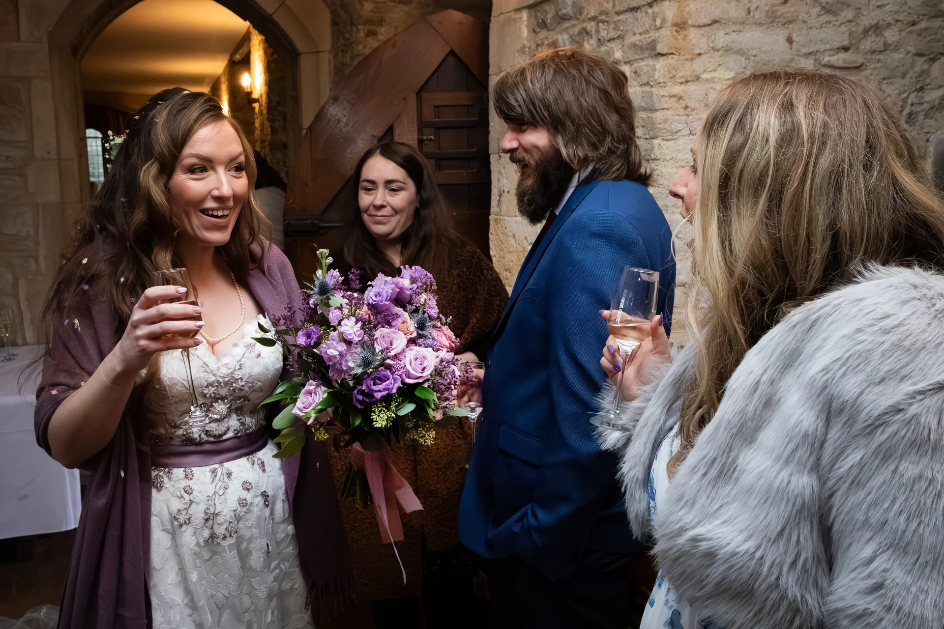 A smiling bride holding a purple floral bouquet and a glass of champagne stands with guests in a rustic stone venue.