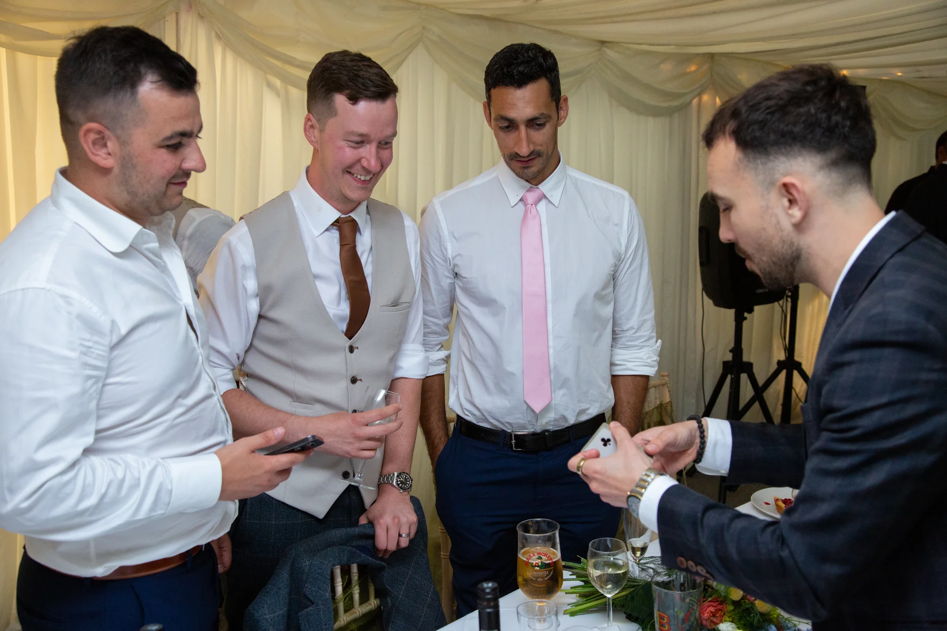 A close-up magic entertainer performs a card trick for a group of smiling groomsmen inside a marquee during a wedding reception.